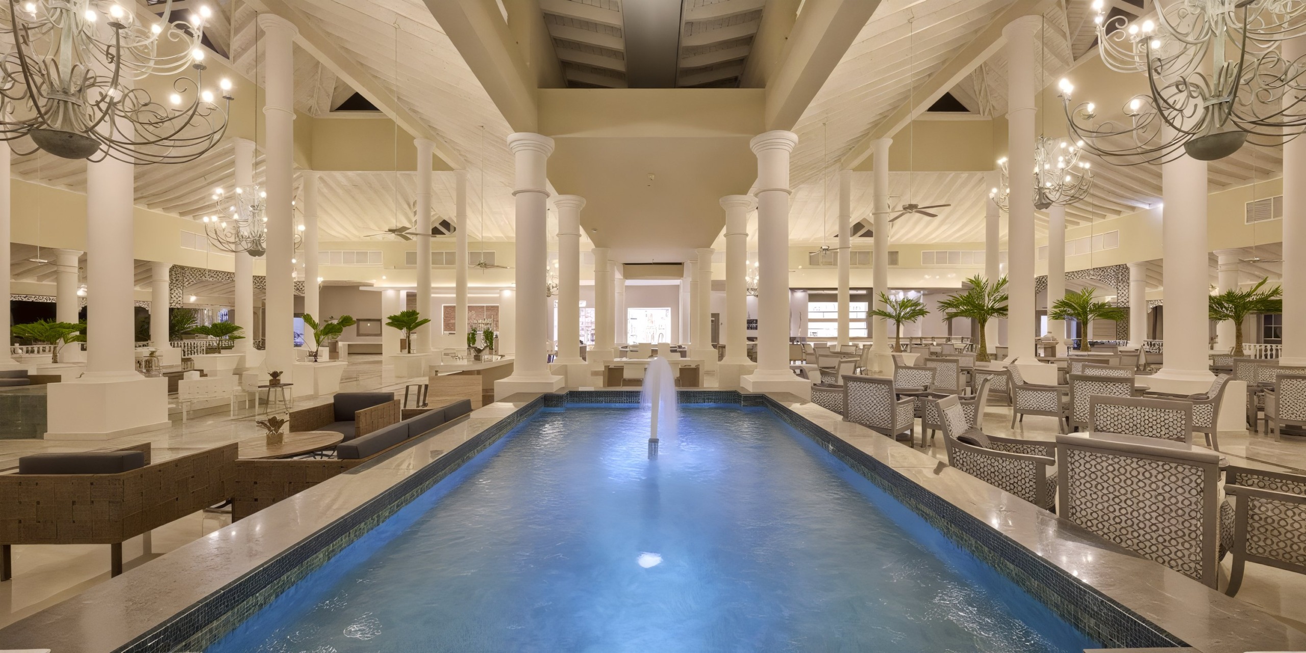 Experience the opulence of Bahia Principe Luxury Ambar with its high ceilings, grand chandeliers, elegant white columns, and a central rectangular fountain surrounded by inviting seating areas.