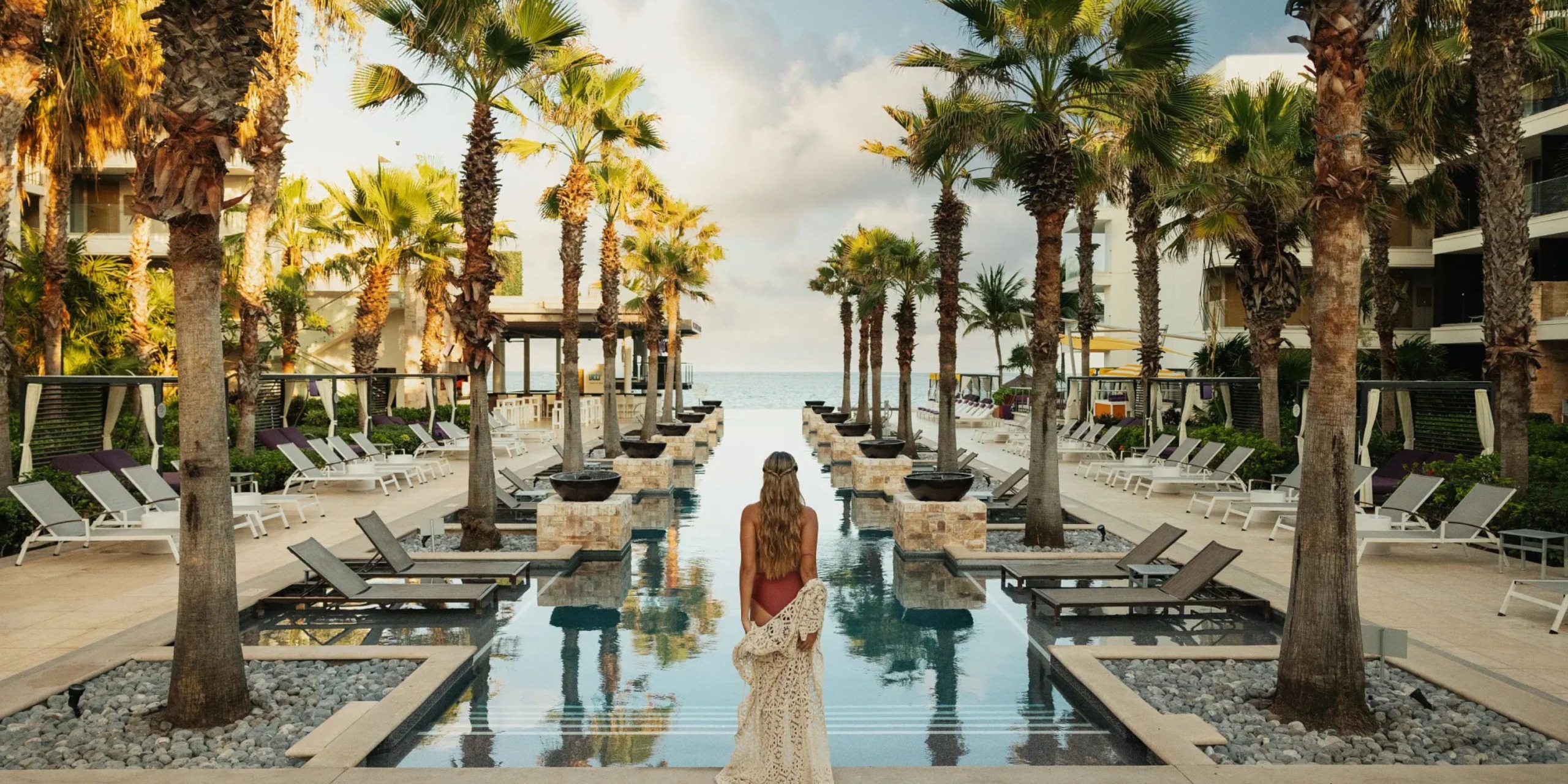 A woman dressed elegantly strolls towards a reflecting pool bordered by palm trees and lounge chairs, all leading to a breathtaking ocean view at the Breathless Riviera Cancún Resort and Spa.