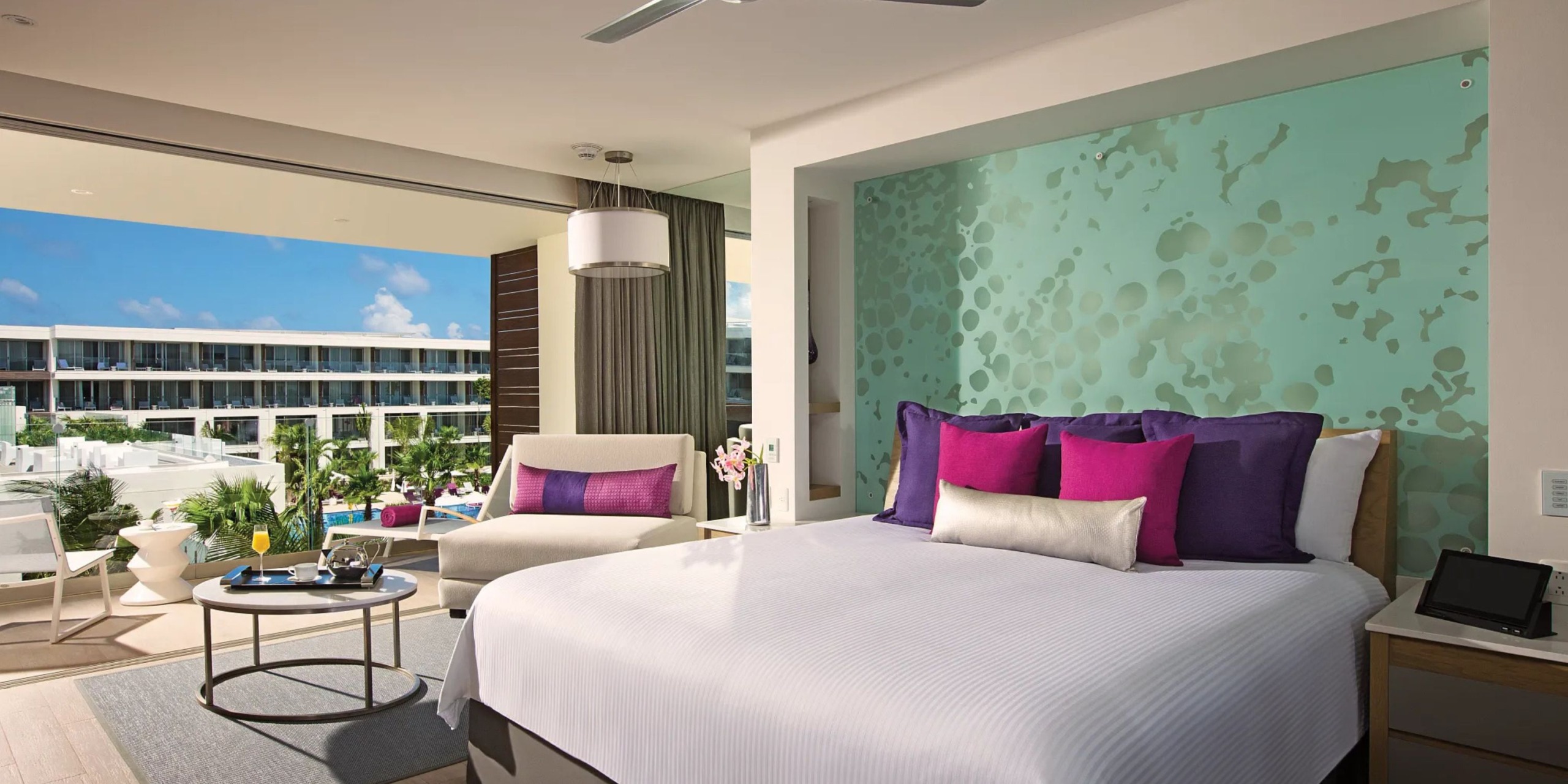 A contemporary room at the Breathless Riviera Cancún Resort and Spa features a comfortable bed with vibrant pillows, a cozy seating area, and a balcony offering views of the resort complete with a pool and lush greenery.