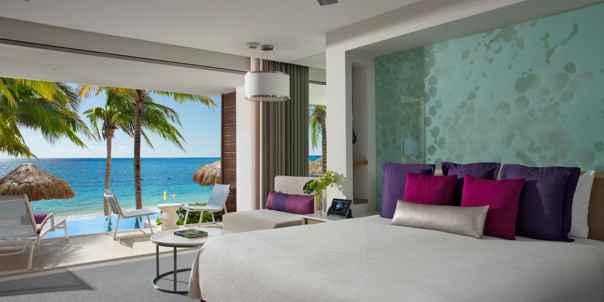 Experience a room at the Breathless Riviera Cancún Resort and Spa, featuring open sliding doors that face the beach. Inside, you'll find a comfortable bed, a cozy seating area, and decor in elegant white and purple tones, all offering stunning views of palm trees and the ocean.