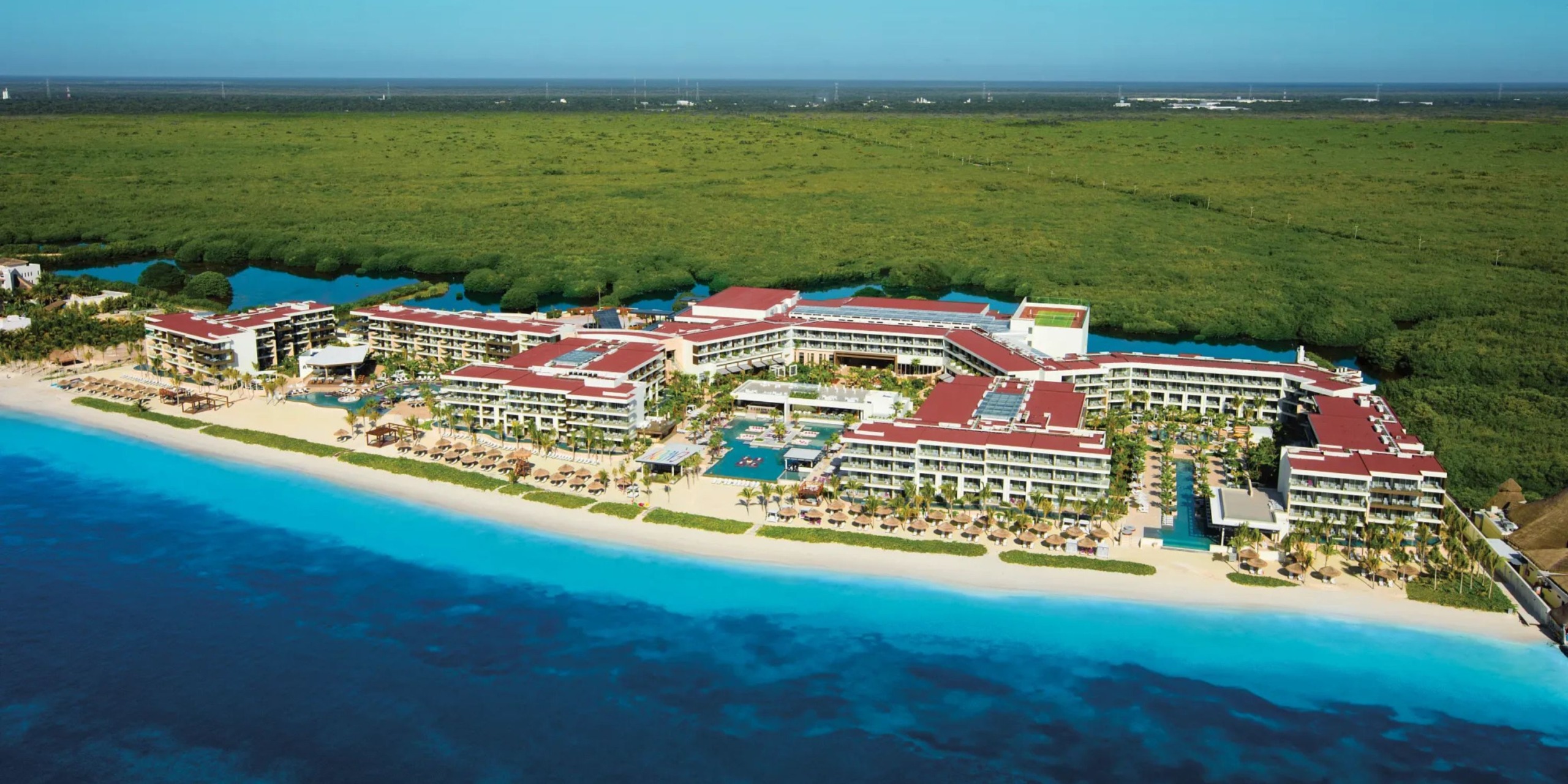 Bird's-eye view of the expansive Breathless Riviera Cancún Resort and Spa, featuring red-roofed buildings amidst lush greenery and clear blue waters.