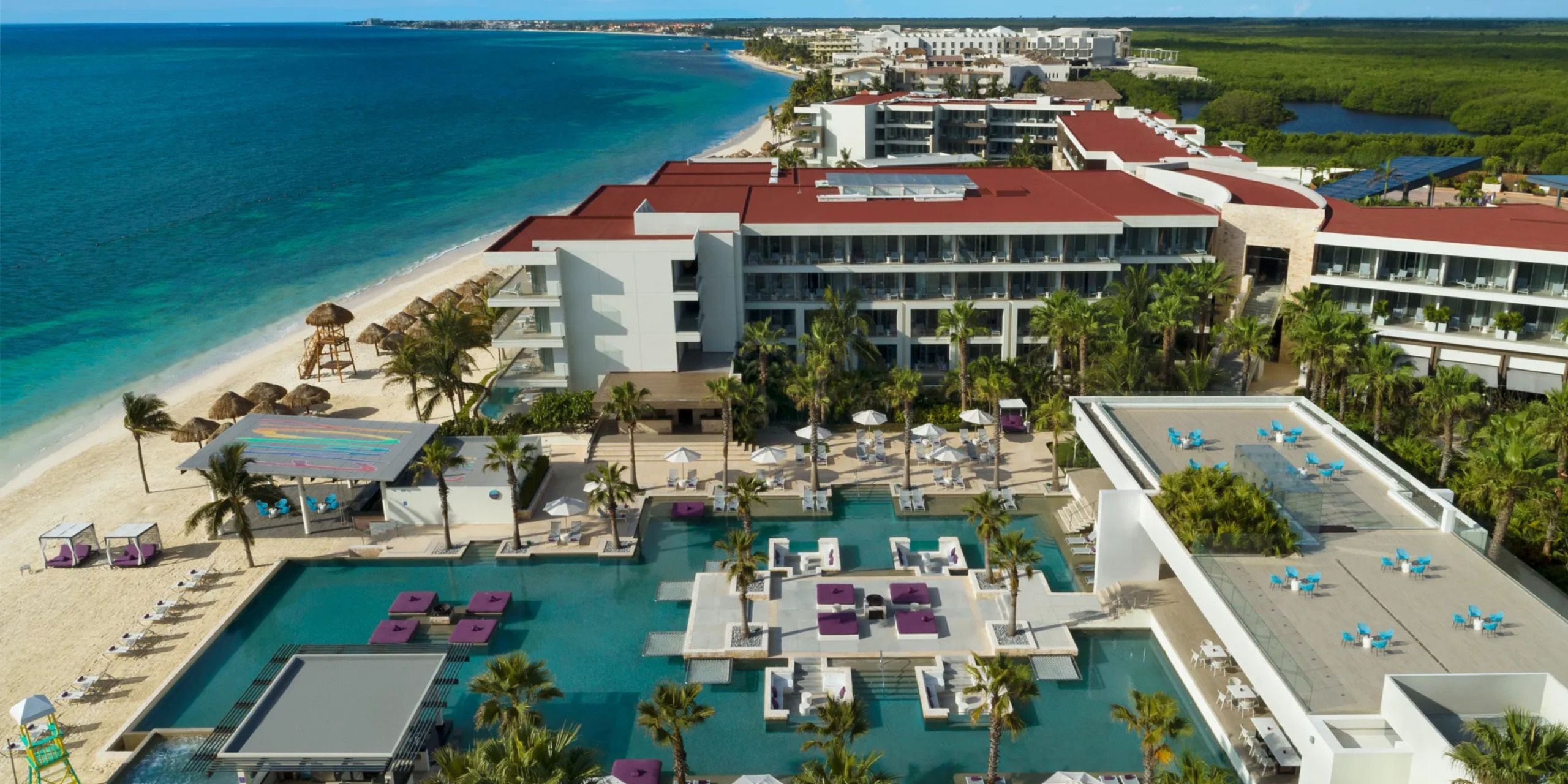 Experience an aerial view of the luxurious Breathless Riviera Cancún Resort and Spa, featuring multiple pools, swaying palm trees, and a pristine white sandy beach bordering the vibrant blue ocean. The resort's red-roofed buildings are charmingly nestled amidst lush vegetation.