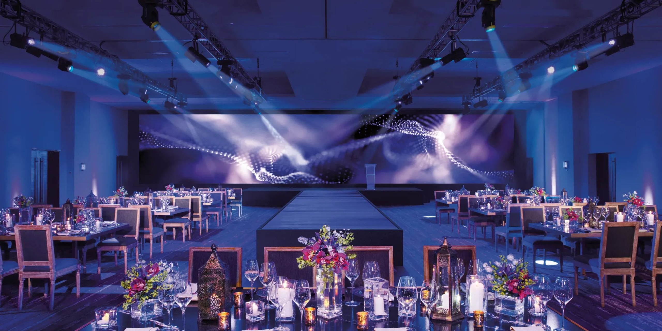 Breathless Riviera Cancún Resort and Spa features a banquet hall equipped with a runway stage, elegantly decorated tables, stunning floral centerpieces, and sophisticated mood lighting.