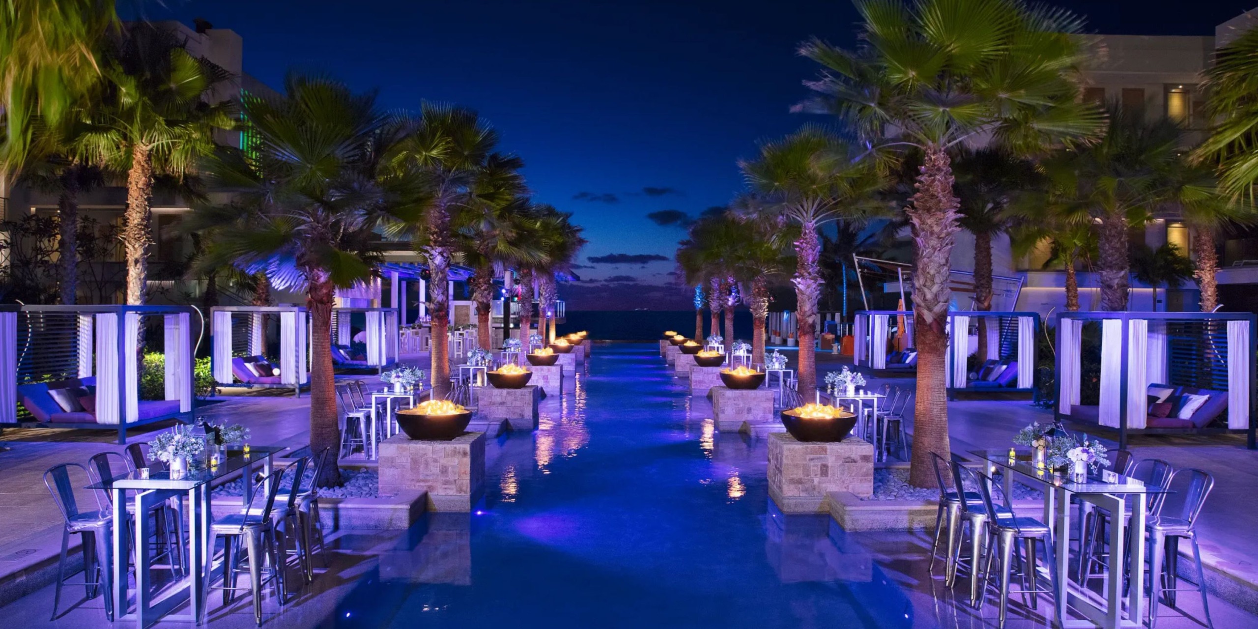 At the Breathless Riviera Cancún Resort and Spa, the outdoor pool area exudes luxury at night, featuring palm trees, cabanas, tables, and glowing fire pits lining the water's edge. The sky is a deep blue with clouds lingering on the horizon.