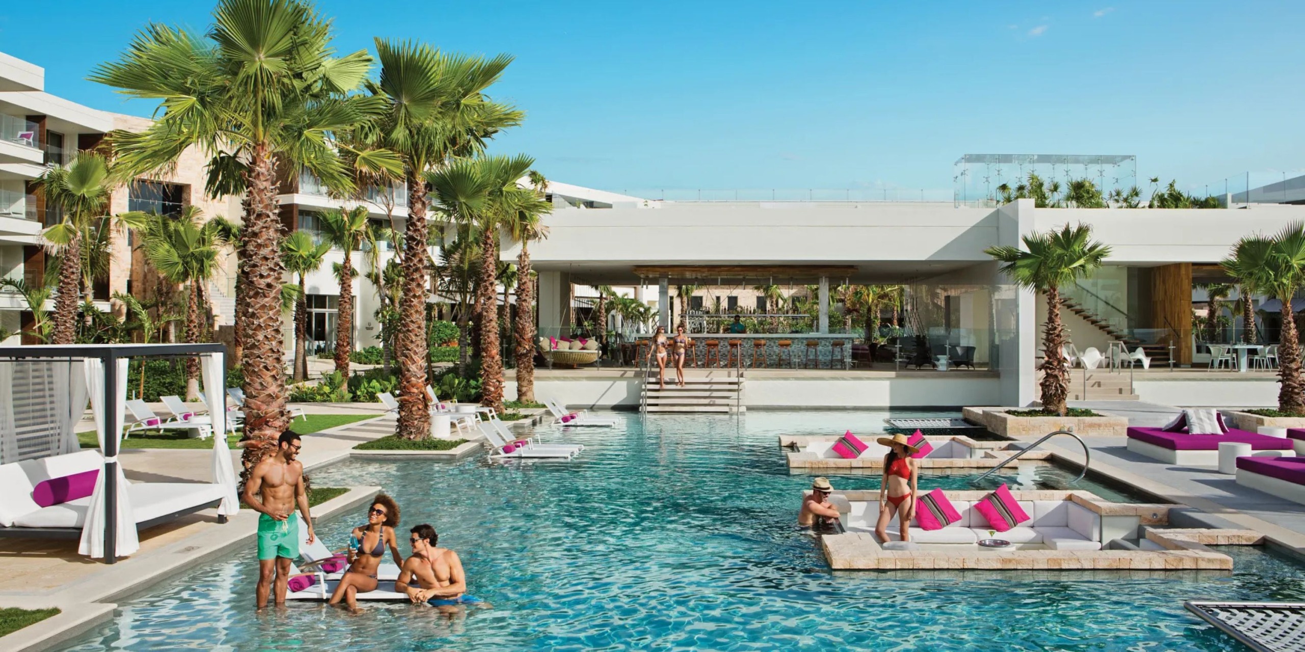 The Breathless Riviera Cancún Resort and Spa features a vibrant pool area where guests can relax and socialize. The surroundings are adorned with palm trees and white buildings, complemented by loungers with pink cushions scattered nearby.