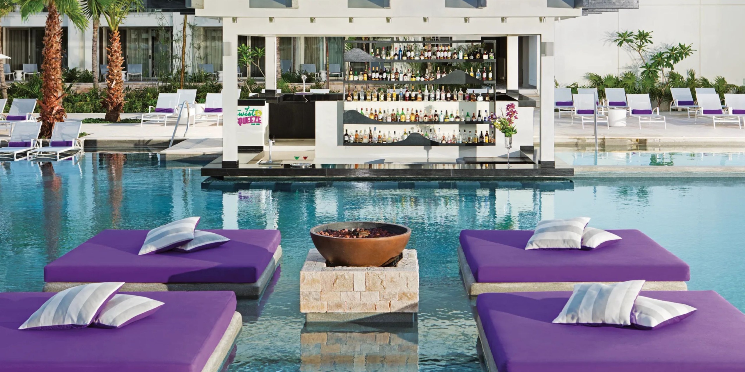 Breathless Riviera Cancún Resort and Spa features purple pool lounge beds with white pillows encircling a round fire pit, while a poolside bar offering a variety of drinks serves as the backdrop.