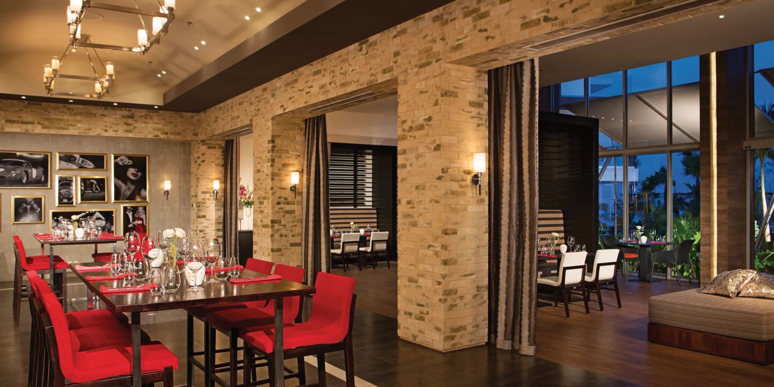 The dining area at Breathless Riviera Cancún Resort and Spa boasts red chairs and elegantly set tables, with brick walls, modern lighting, and large windows that offer a stunning night view.