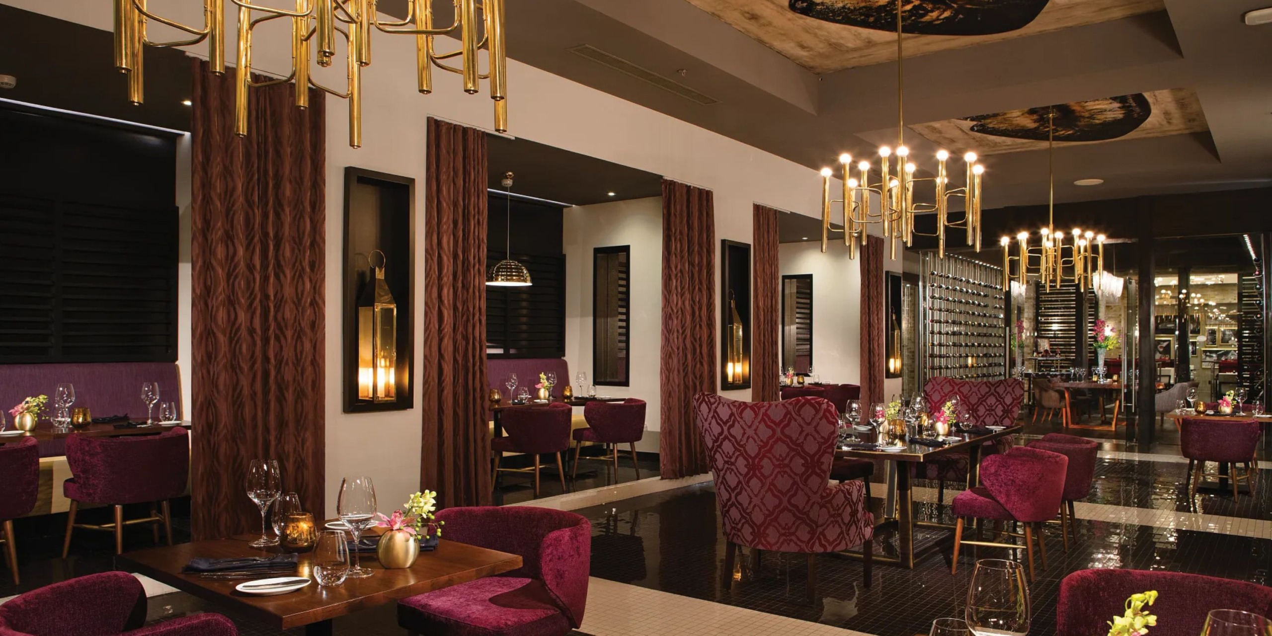 The interior of the Breathless Riviera Cancún Resort and Spa features an elegant restaurant adorned with red chairs, dark wood tables, and decorative chandeliers. Mirrors and ornate curtains embellish the walls, while dim lighting creates a cozy ambiance.