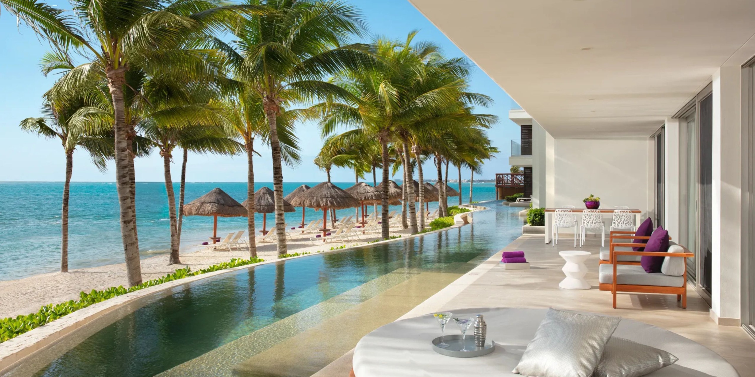 Enjoy a beachfront view from the modern terrace at Breathless Riviera Cancún Resort and Spa, featuring palm trees, lounge chairs, a long pool, and shaded cabanas overlooking the ocean.