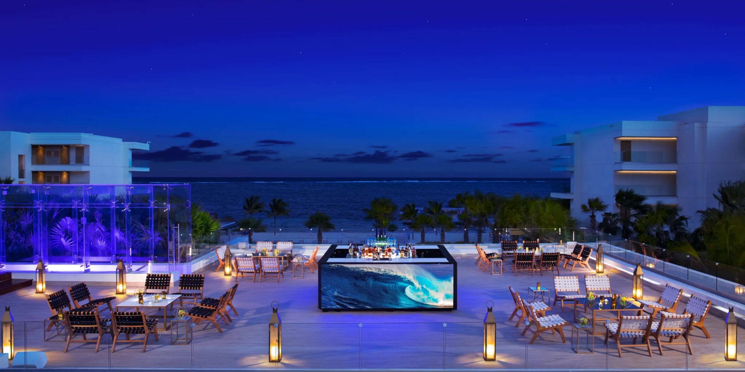 Experience the mesmerizing rooftop bar at Breathless Riviera Cancún Resort and Spa, where you'll enjoy a nighttime ocean view surrounded by glowing lanterns. The ambiance features a central video screen showcasing a wave, with tables and chairs thoughtfully arranged throughout the space.