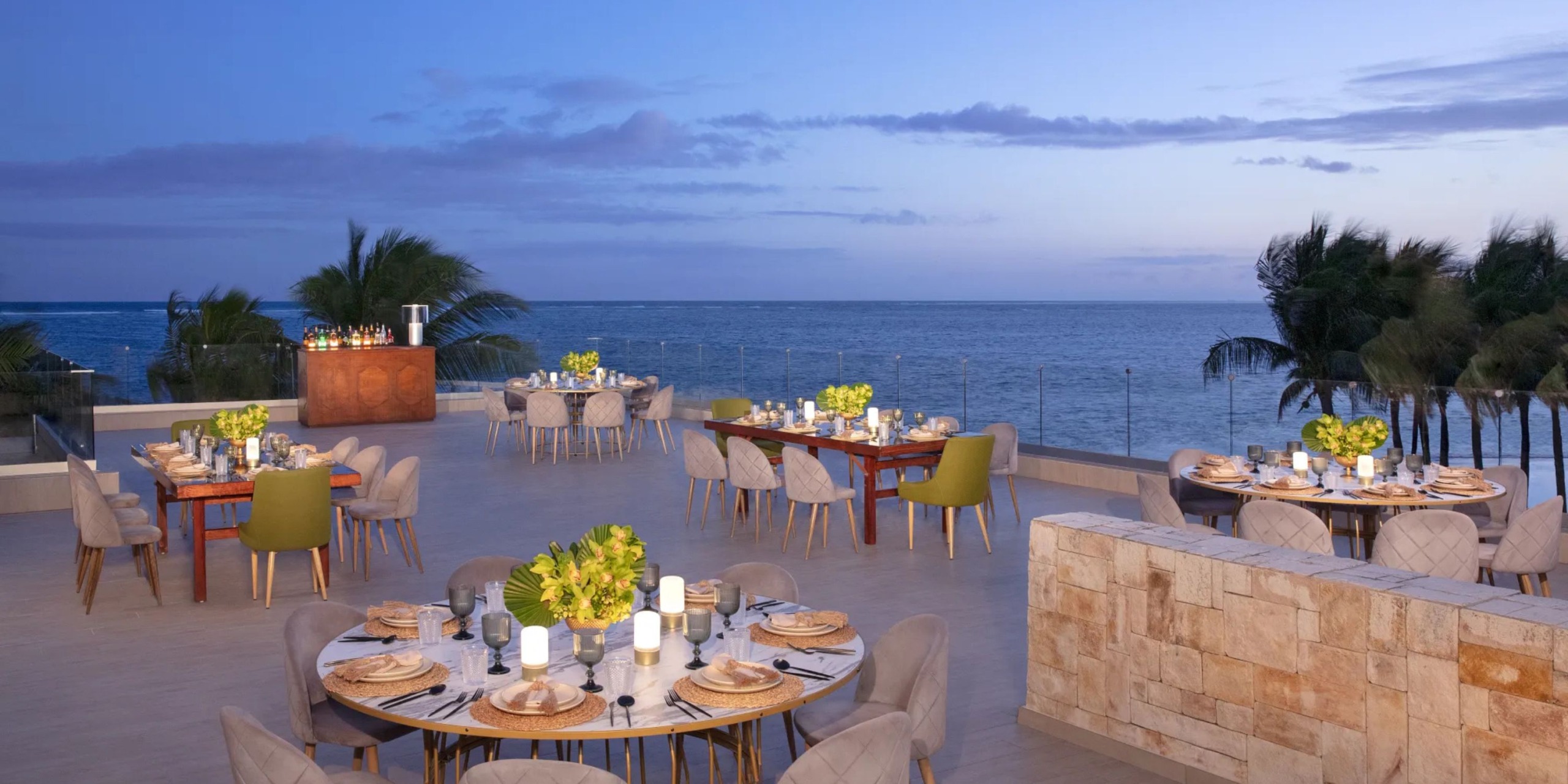 At the Breathless Riviera Cancún Resort and Spa, enjoy an outdoor dining area with round tables set for meals, offering a stunning ocean view at dusk. Palm trees provide a picturesque backdrop, complemented by a bar at one end.