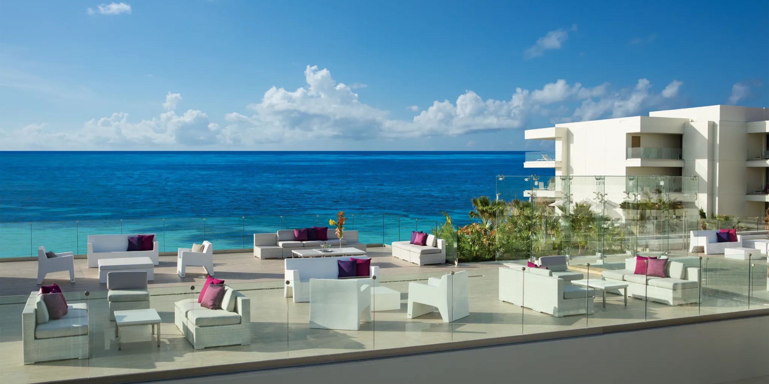The patio at Breathless Riviera Cancún Resort and Spa features white furniture with purple cushions, providing a stunning view of the blue ocean and clear sky, next to a modern white building.