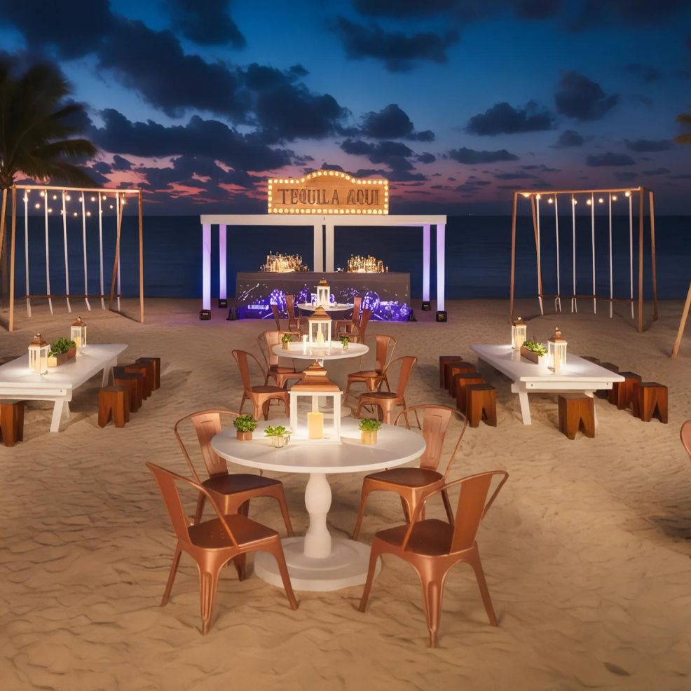 Outdoor beach event setup at sunset with tables and chairs arranged on the sand, a “Tequila Aqui” sign illuminated above a bar, and string lights creating a festive ambiance.