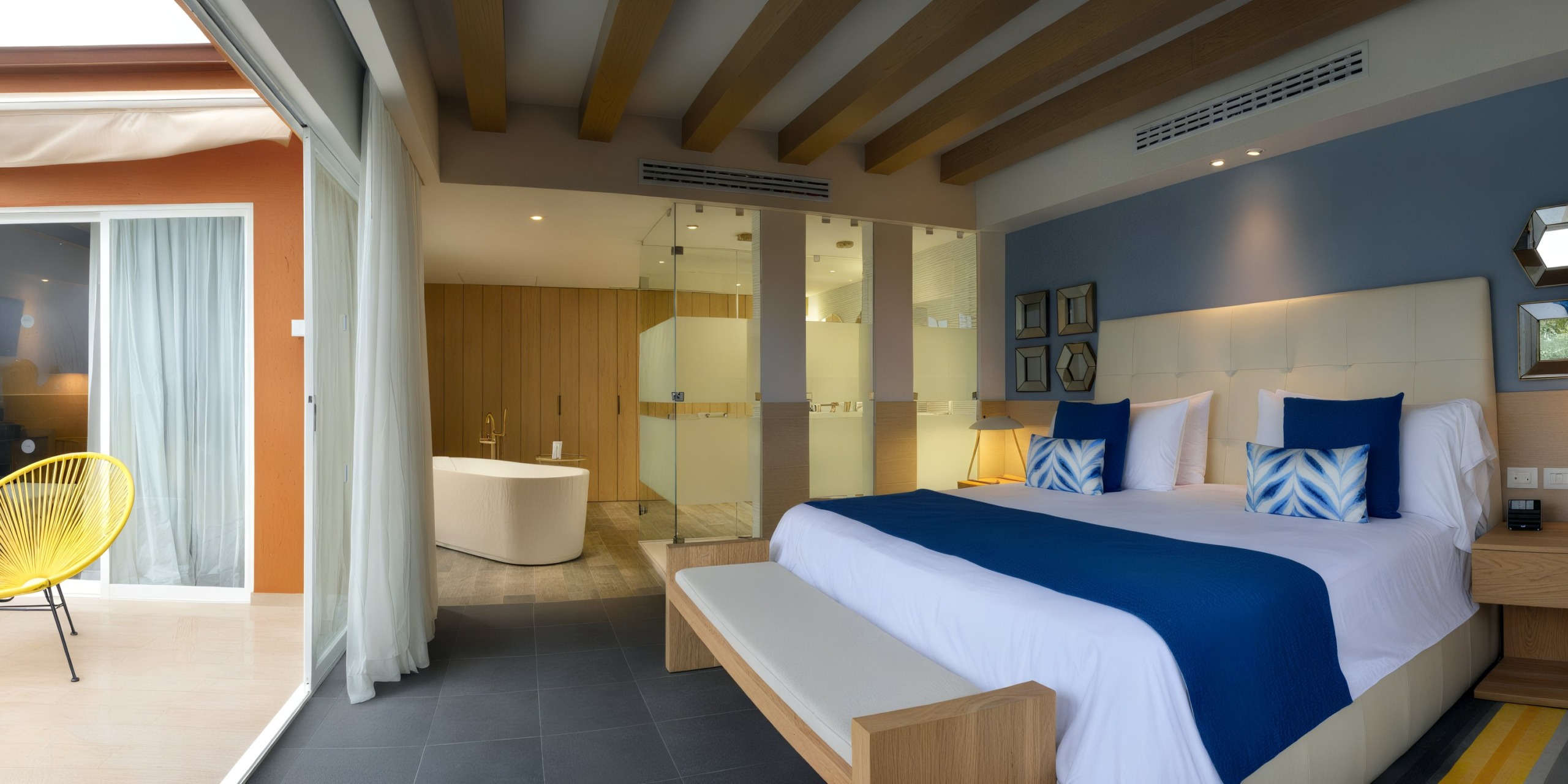 Contemporary hotel room at Catalonia Playa Maroma featuring a spacious bed, blue details, and a bathroom enclosed with glass walls. A small balcony with a yellow chair can be seen through the sliding glass doors.