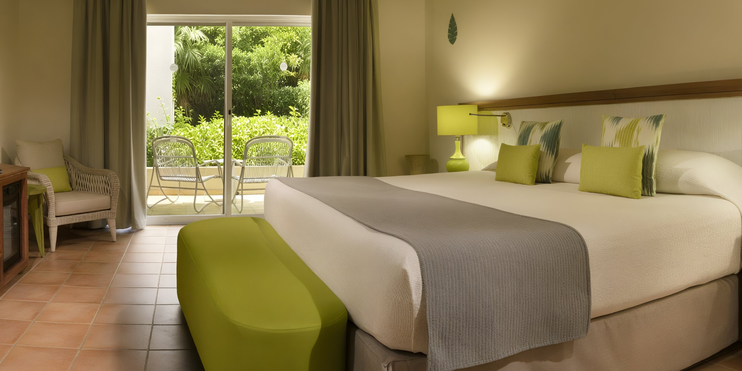 The Catalonia Playa Maroma features a hotel room with a king-sized bed, complemented by green cushions and a grey throw. Through the sliding glass doors, you can access a small patio with chairs that overlooks a lush garden.
