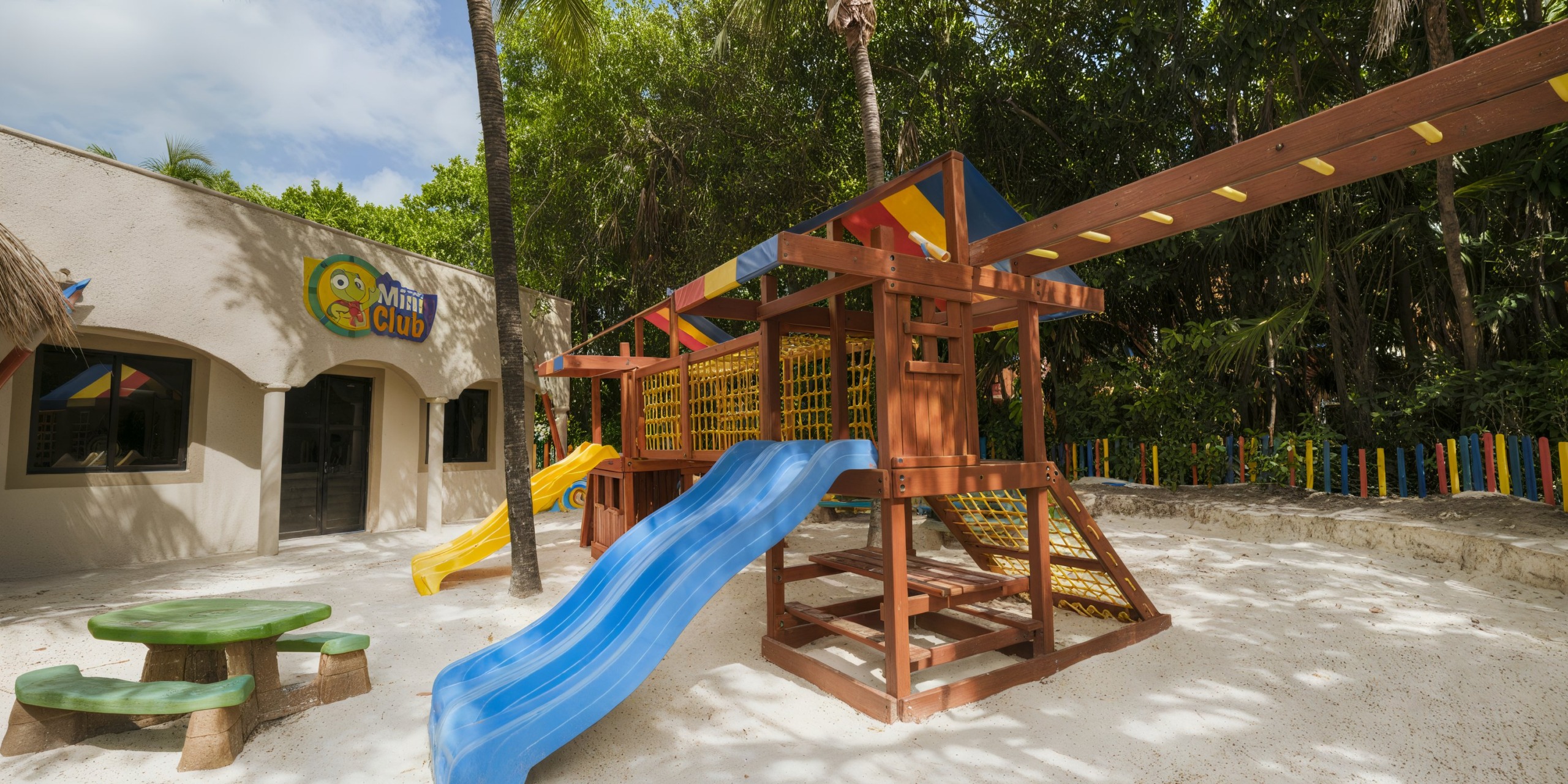 Children's playground featuring a slide and climbing structure, set amidst sand and trees, located near a building labeled "Mini Club" at Catalonia Playa Maroma.