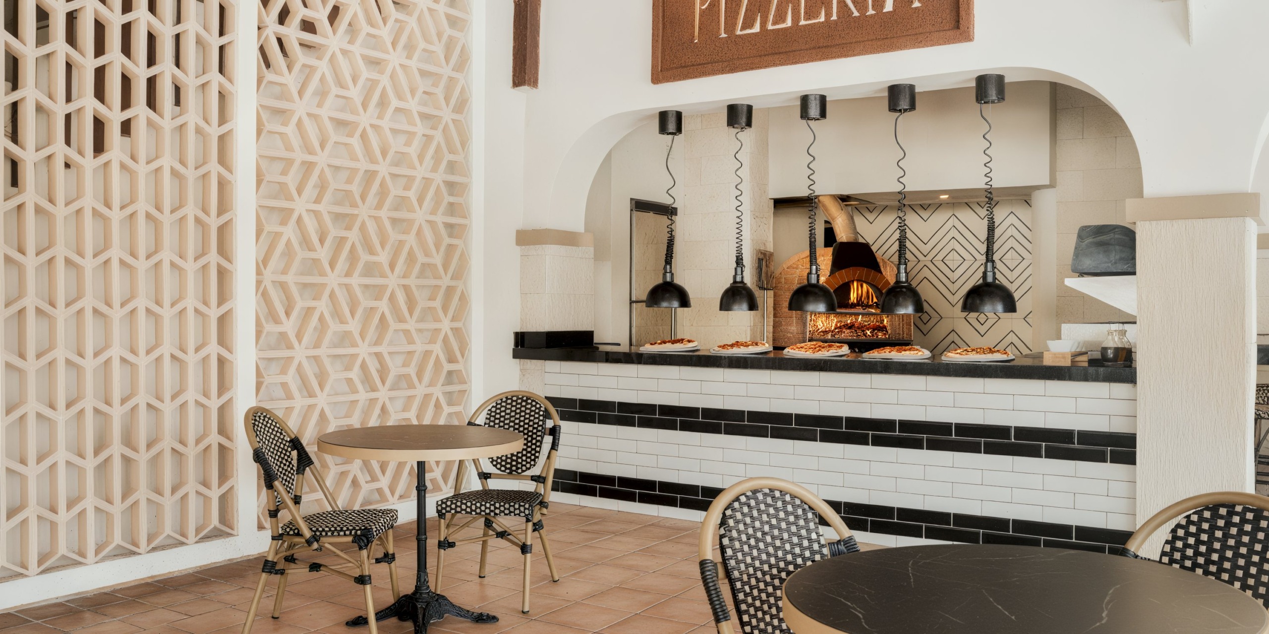 The Catalonia Playa Maroma features a pizzeria with a counter displaying pizzas, pendant lights, and a wood-fired oven. In the foreground, there are two round tables accompanied by chairs.