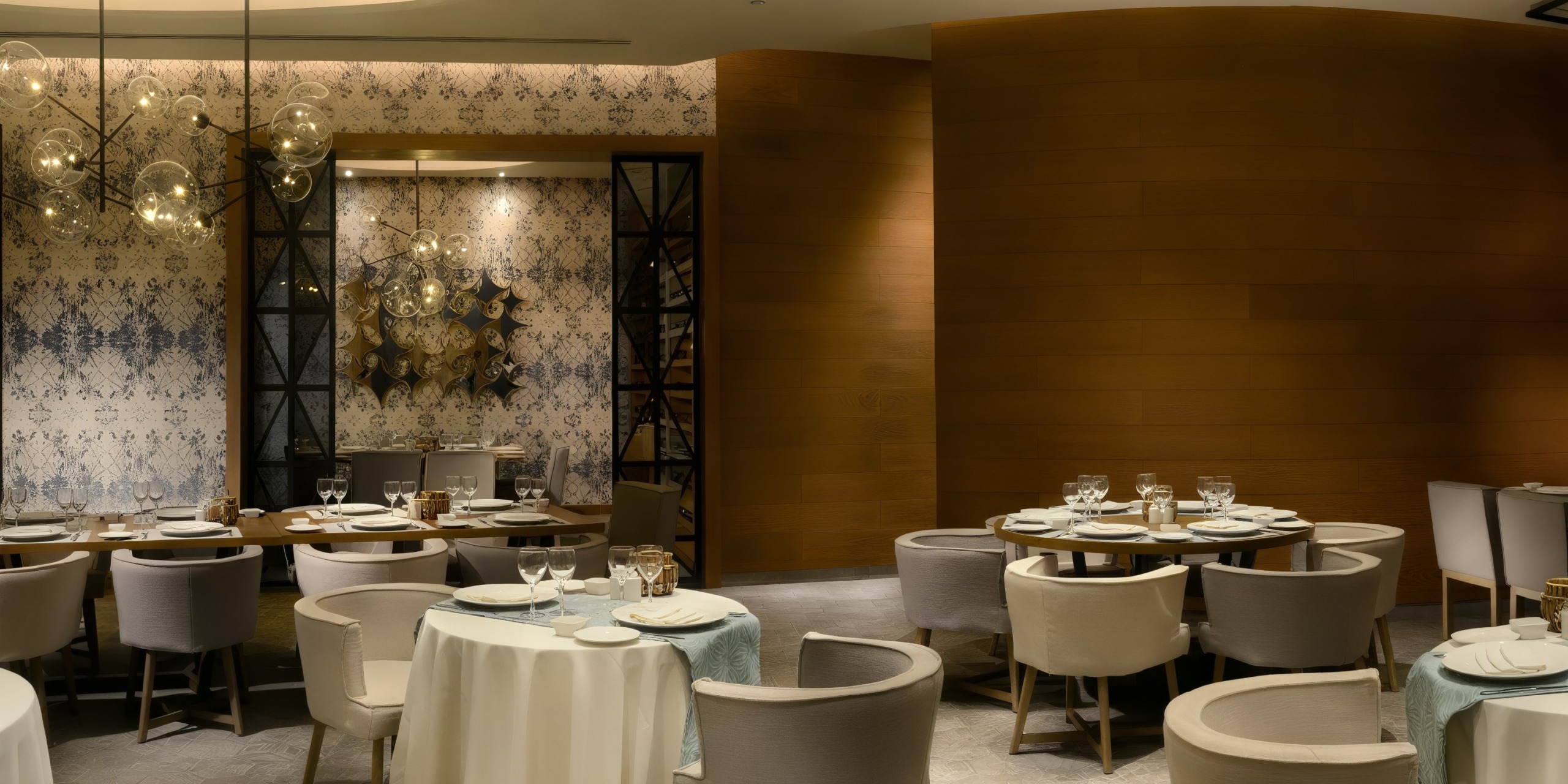 The Catalonia Playa Maroma features a contemporary restaurant interior with round tables draped in white tablecloths, encircled by light gray chairs. The space is elegantly enhanced with modern lighting and decorative wall patterns.