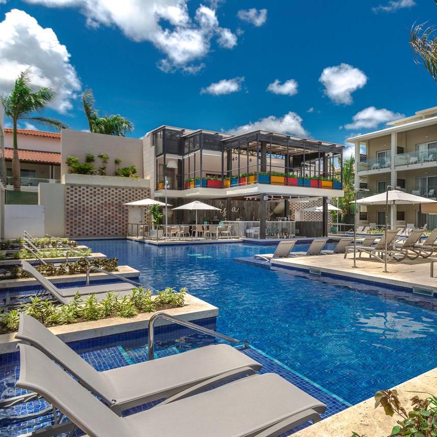 A luxury Catalonia Royal La Romana pool with lounge chairs, ideal for a destination wedding, surrounded by modern buildings under a clear blue sky.