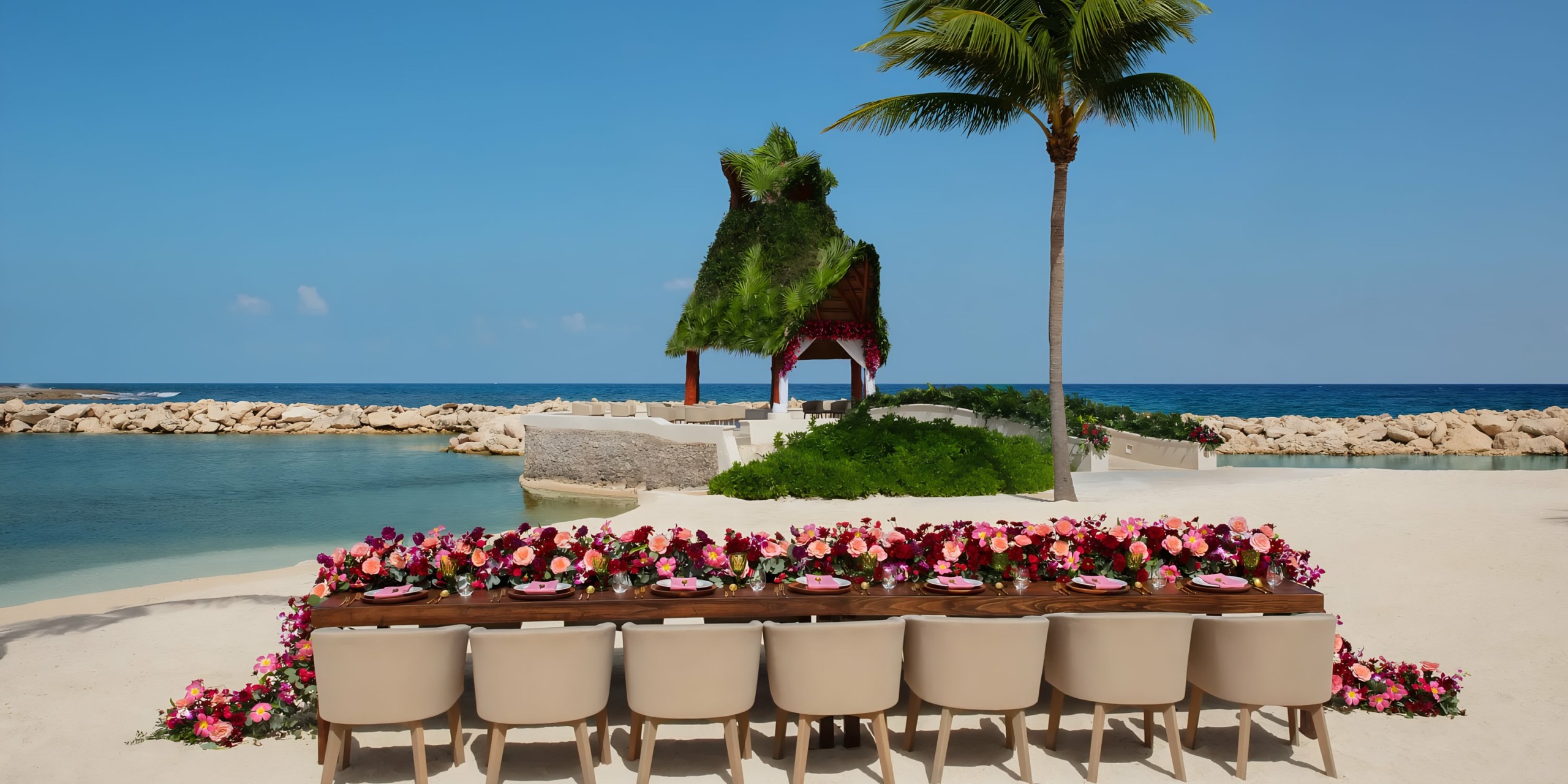 Outdoor dining setup by the beach with a long table set for a meal and a palm-fringed Dreams Aventuras Riviera Maya in the background.