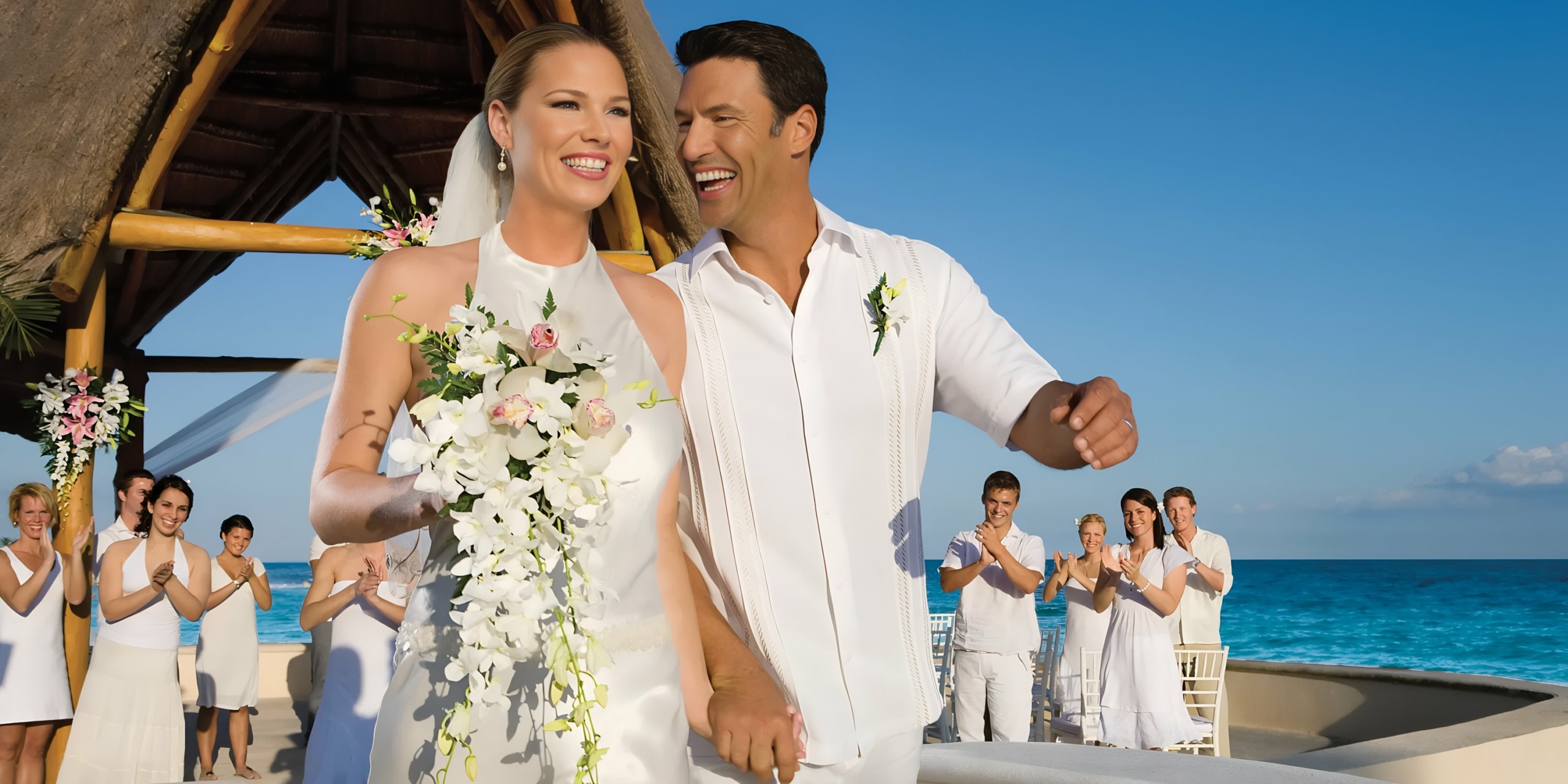 A smiling couple in wedding attire walking hand in hand with applauding guests in the background at a beachside Dreams Aventuras Riviera Maya ceremony.