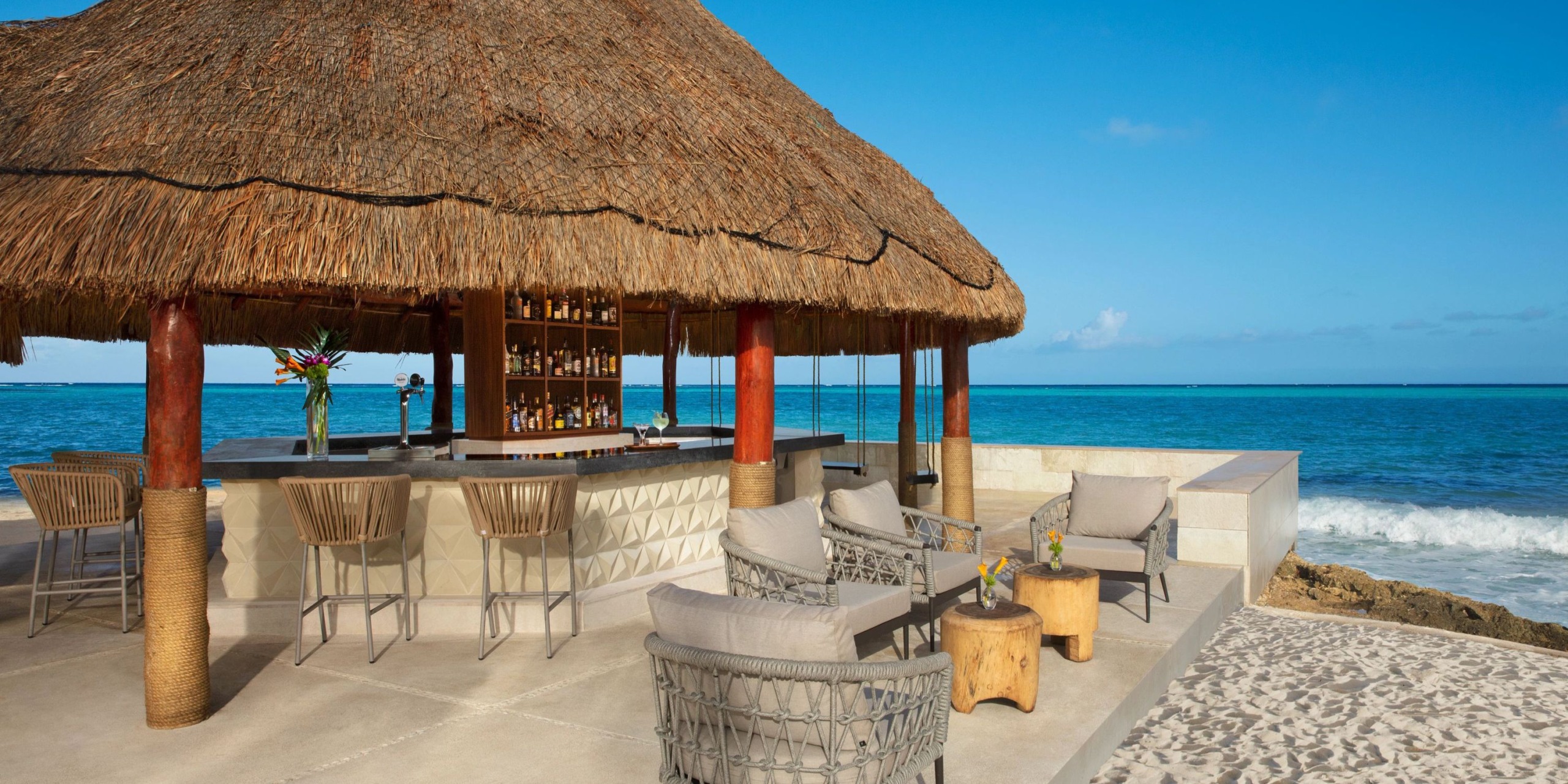 At Dreams Aventuras Riviera Maya, enjoy an outdoor beach bar featuring a thatched roof, surrounded by comfortable stools and lounge chairs with a stunning view of the ocean beneath a crystal-clear blue sky.