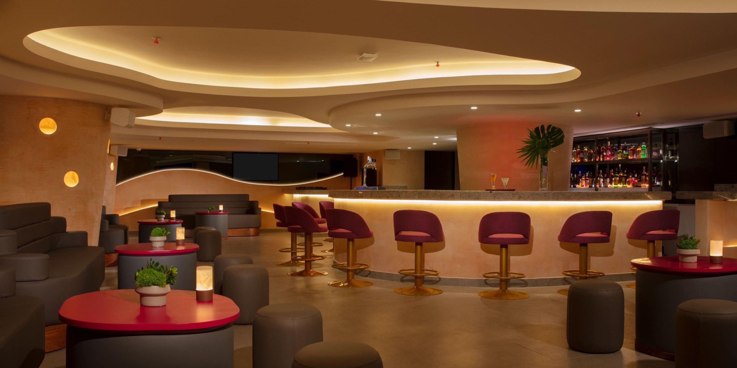 Dreams Aventuras Riviera Maya boasts a contemporary bar interior featuring a curved ceiling, purple bar stools, and ambient lighting. The space is elegantly arranged with tables adorned with small plants and candles scattered throughout.
