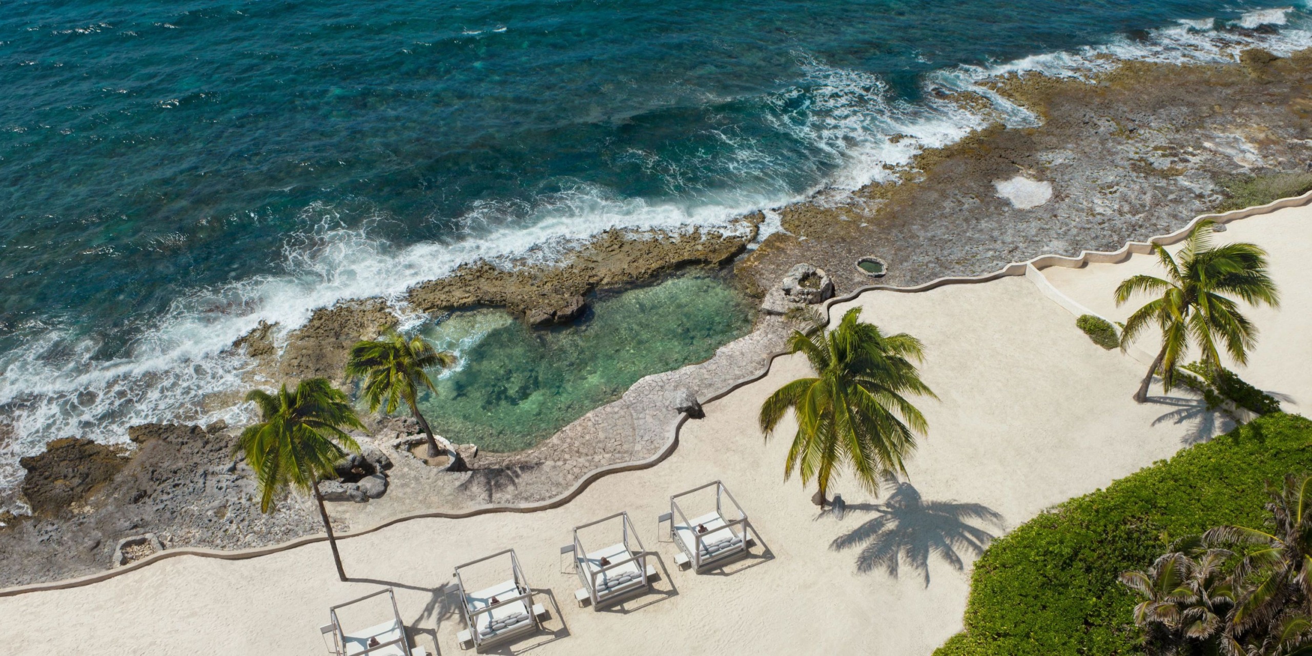 An aerial view showcases the Dreams Aventuras Riviera Maya, featuring a beach lined with four lounge chairs, surrounded by palm trees and a rocky shoreline next to the ocean.