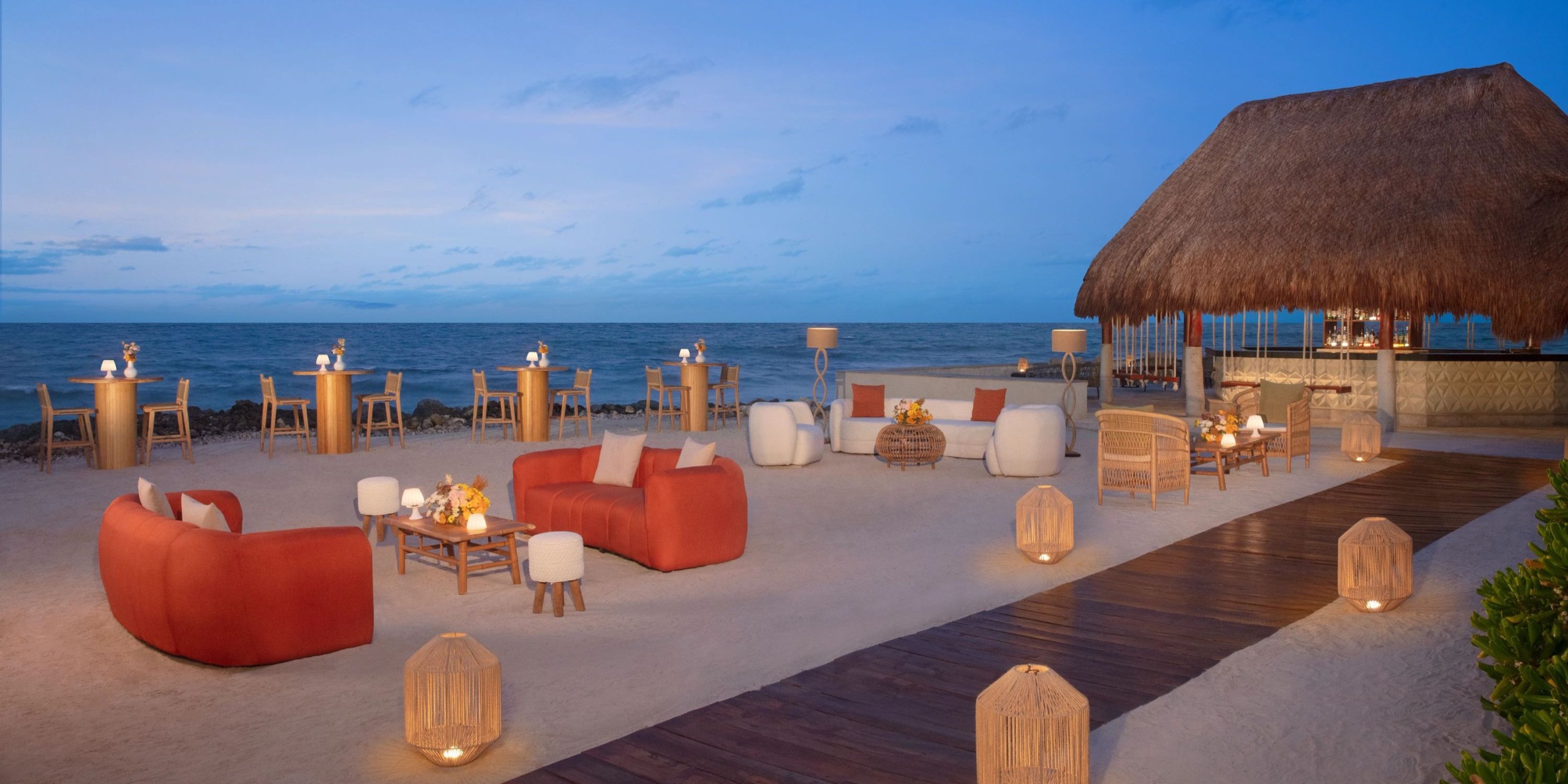 Experience the serene setting at Dreams Aventuras Riviera Maya, featuring red and beige furniture on sandy shores, near a charming thatched-roof structure. Candlelit lanterns illuminate tables arranged along a wooden walkway, with the ocean providing a stunning backdrop.