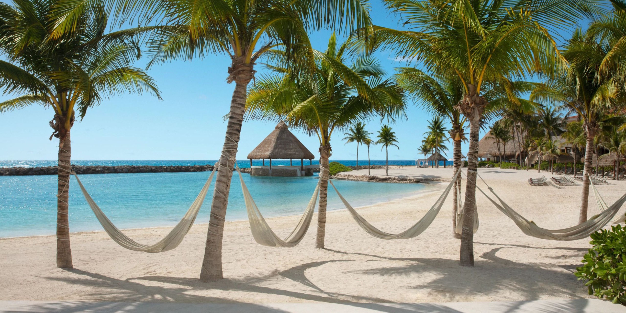 Hammocks are strung between palm trees on the sandy beach at Dreams Aventuras Riviera Maya, with a thatched hut and the calm blue sea in the background.