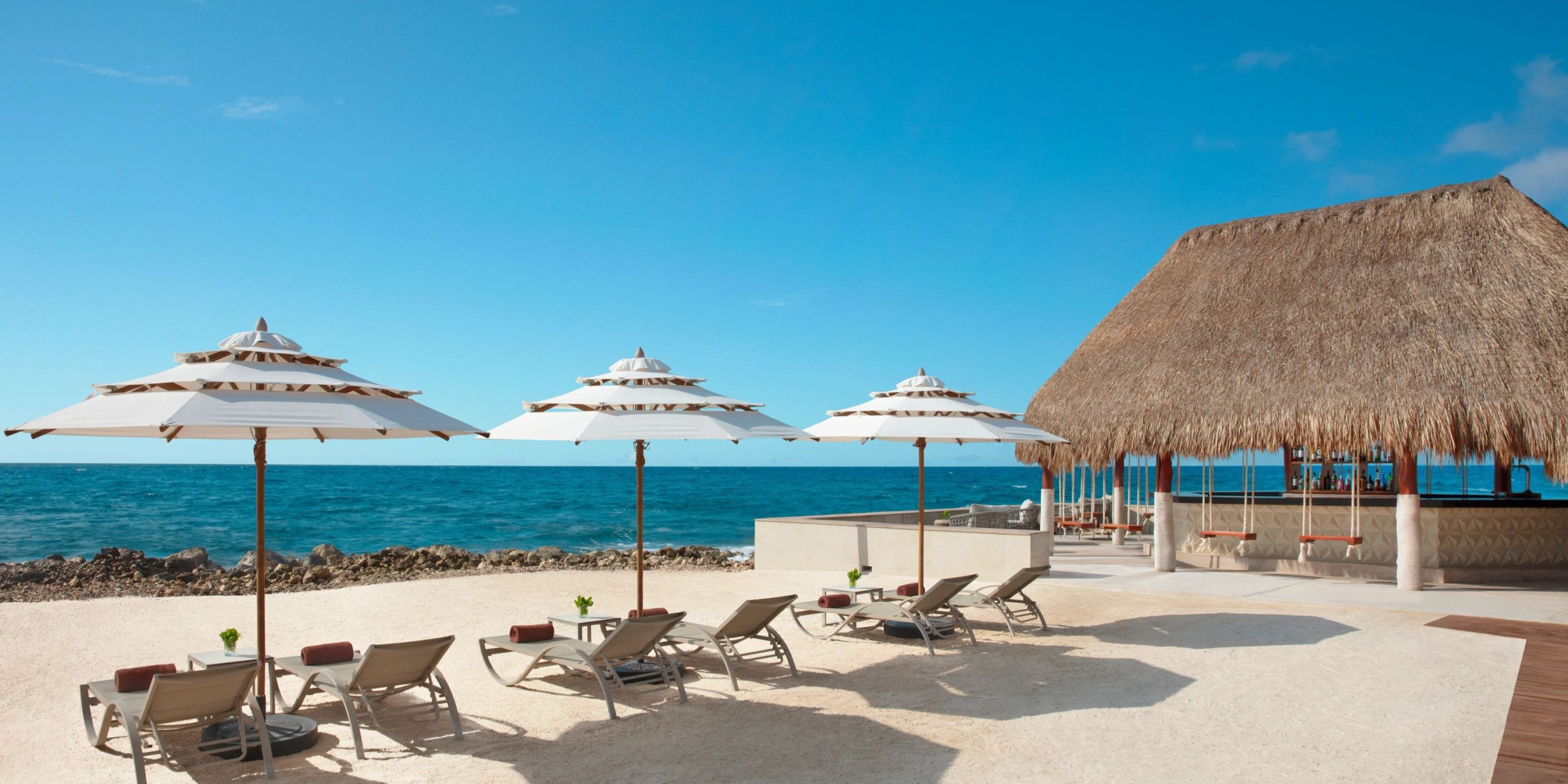 Dreams Aventuras Riviera Maya features a beach scene with lounge chairs under umbrellas near a thatched-roof building, all overlooking the ocean on a clear day.