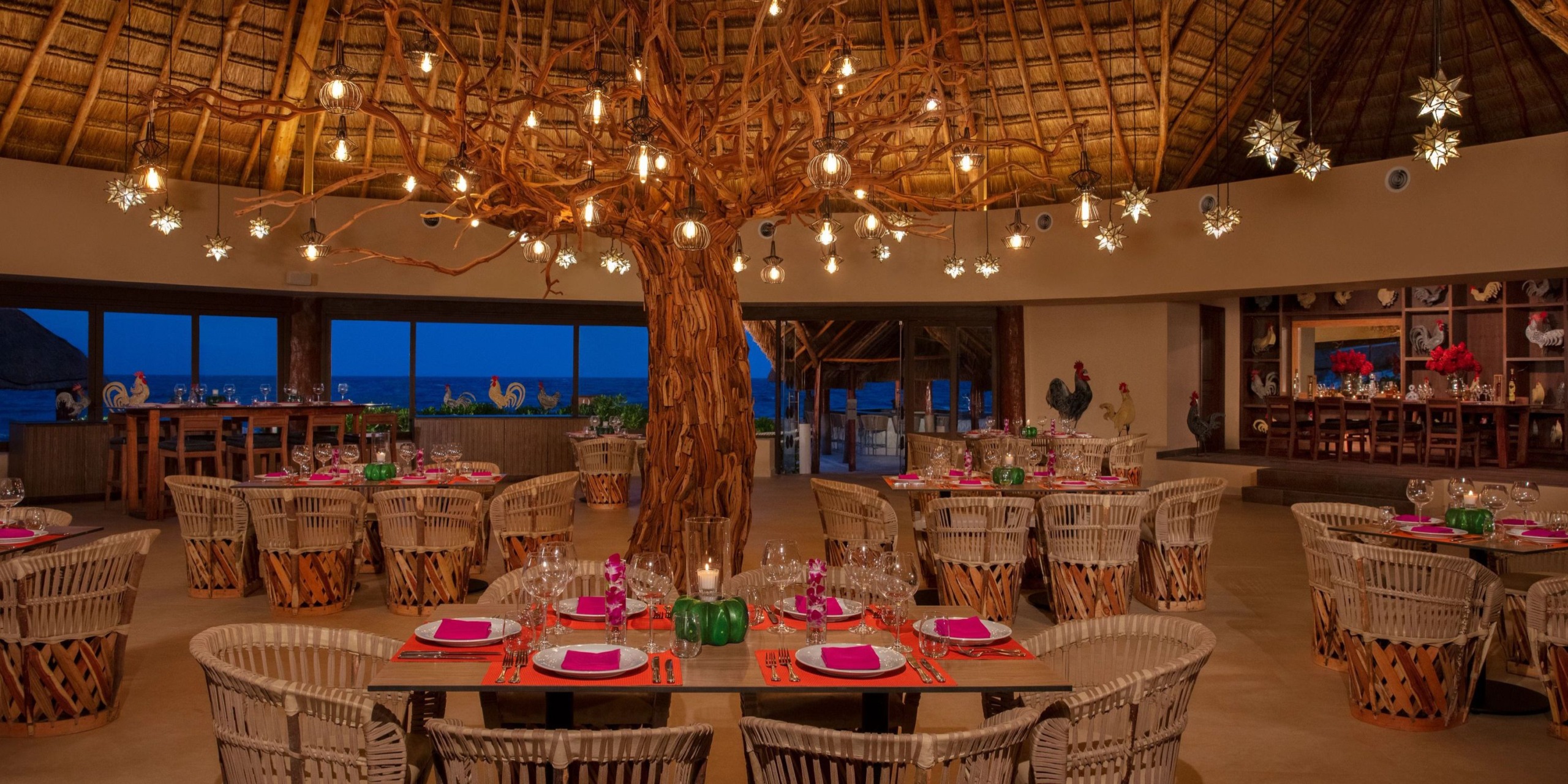 Experience the Dreams Aventuras Riviera Maya restaurant, featuring a tropical-themed interior with a stunning tree sculpture centerpiece. Surrounding the sculpture are woven chairs and tables adorned with pink place settings, all illuminated by decorative overhead lights.