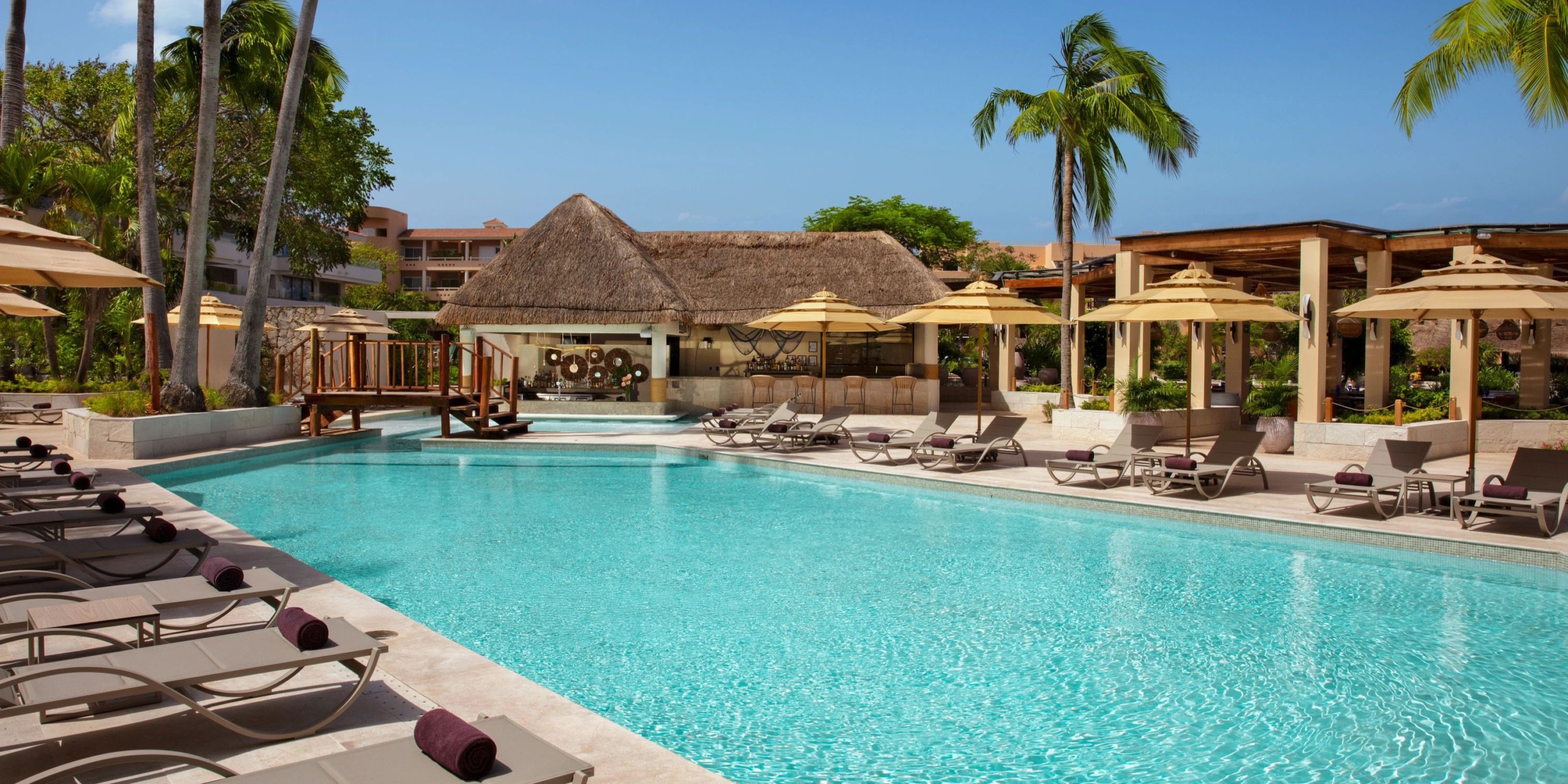 The Dreams Aventuras Riviera Maya features an outdoor swimming pool complete with lounge chairs, umbrellas, and a charming thatched-roof structure, set against a backdrop of palm trees and nearby buildings.