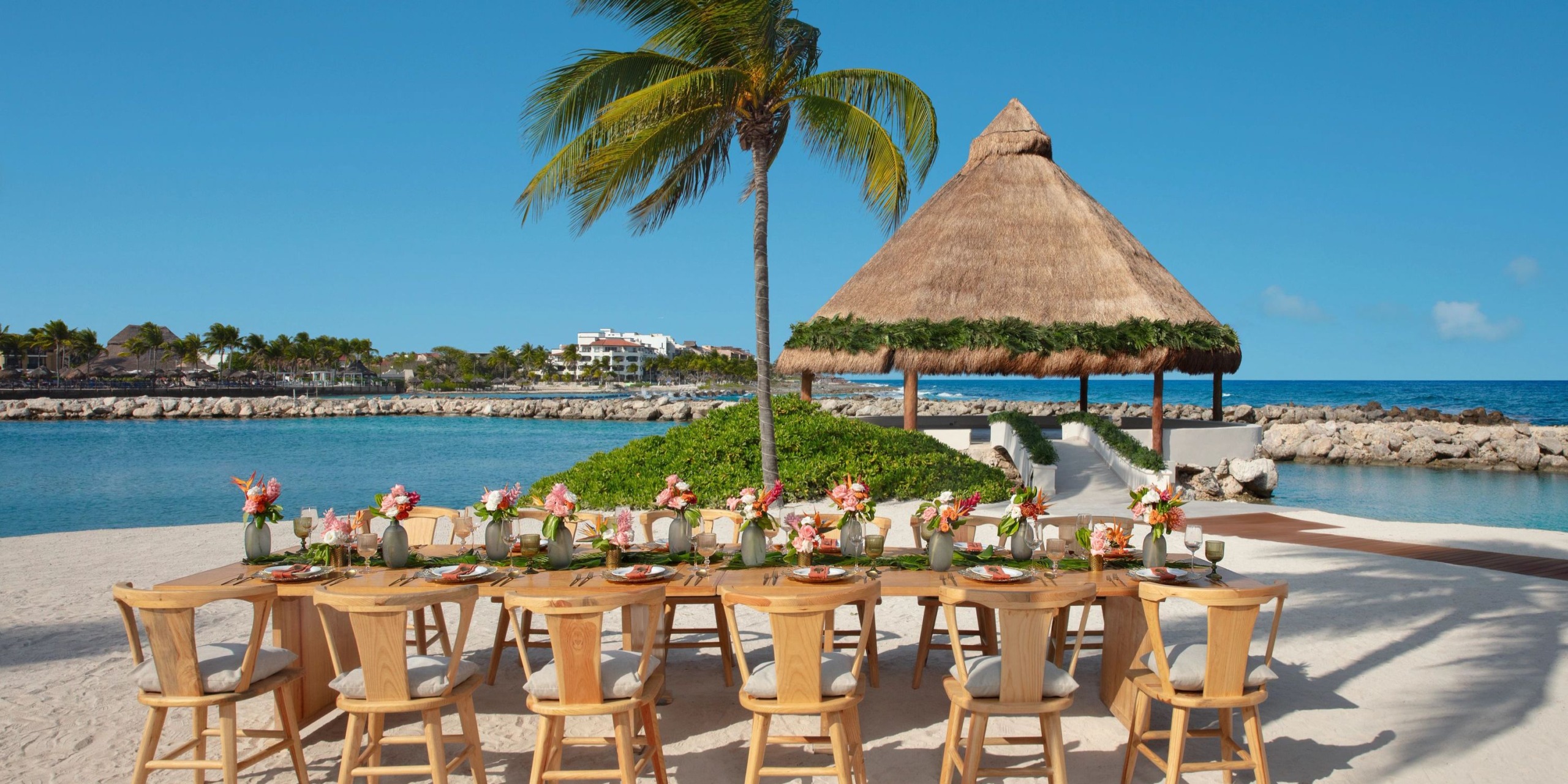 Outdoor dining setup at the Dreams Aventuras Riviera Maya, adorned with floral arrangements, featuring a long wooden table and chairs. A thatched-roof hut and palm tree overlook a calm sea and blue sky.
