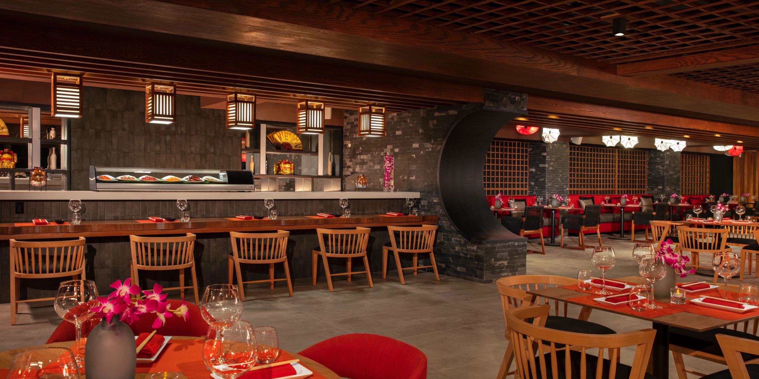 The interior of Dreams Aventuras Riviera Maya features a contemporary design with wooden chairs, red accents, and pendant lights. The tables are elegantly set with glasses and red napkins, while a sushi bar can be seen in the background.