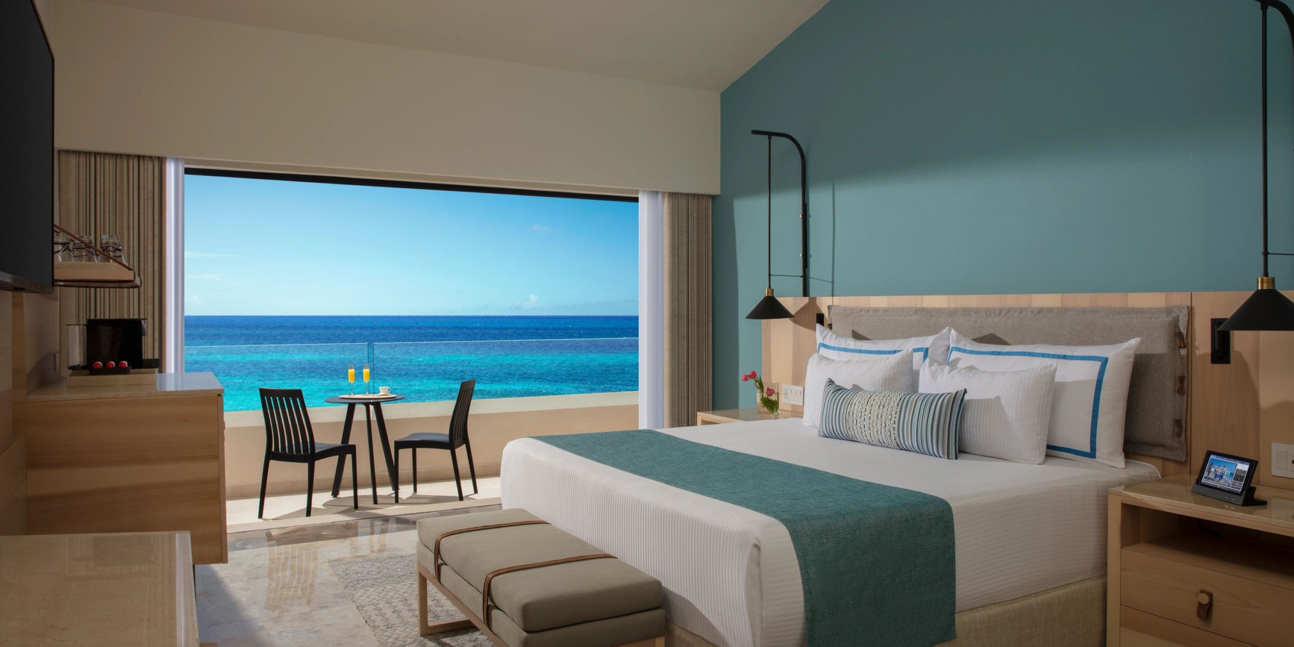 A contemporary room at the Dreams Aventuras Riviera Maya, featuring a spacious bed, bench, and table set by a window with views of the azure ocean and sky.