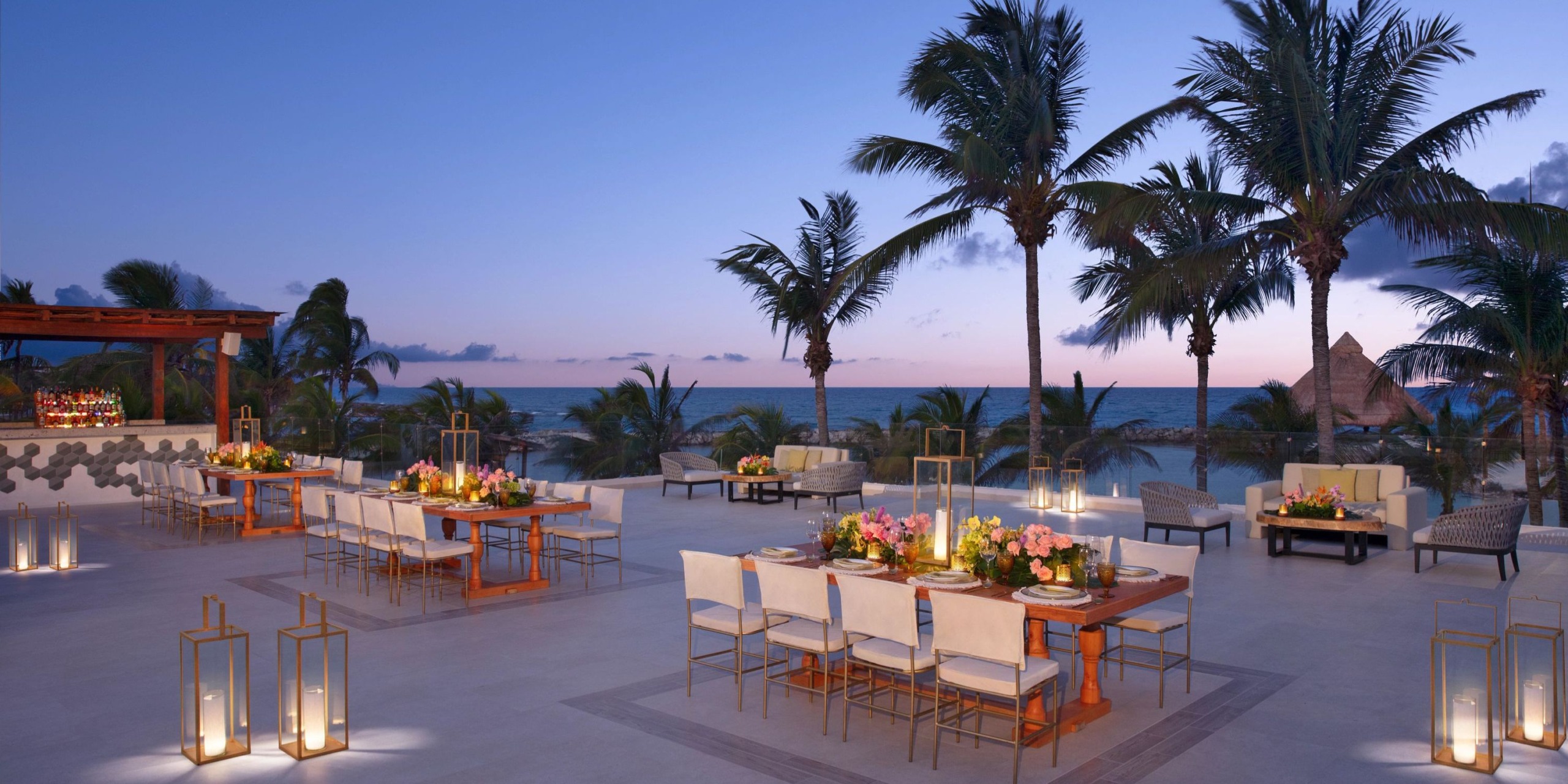 The outdoor dining area at Dreams Aventuras Riviera Maya provides a serene setting with ocean views at sunset, featuring tables adorned with floral centerpieces. The tranquility is further enhanced by palm trees and lanterns.