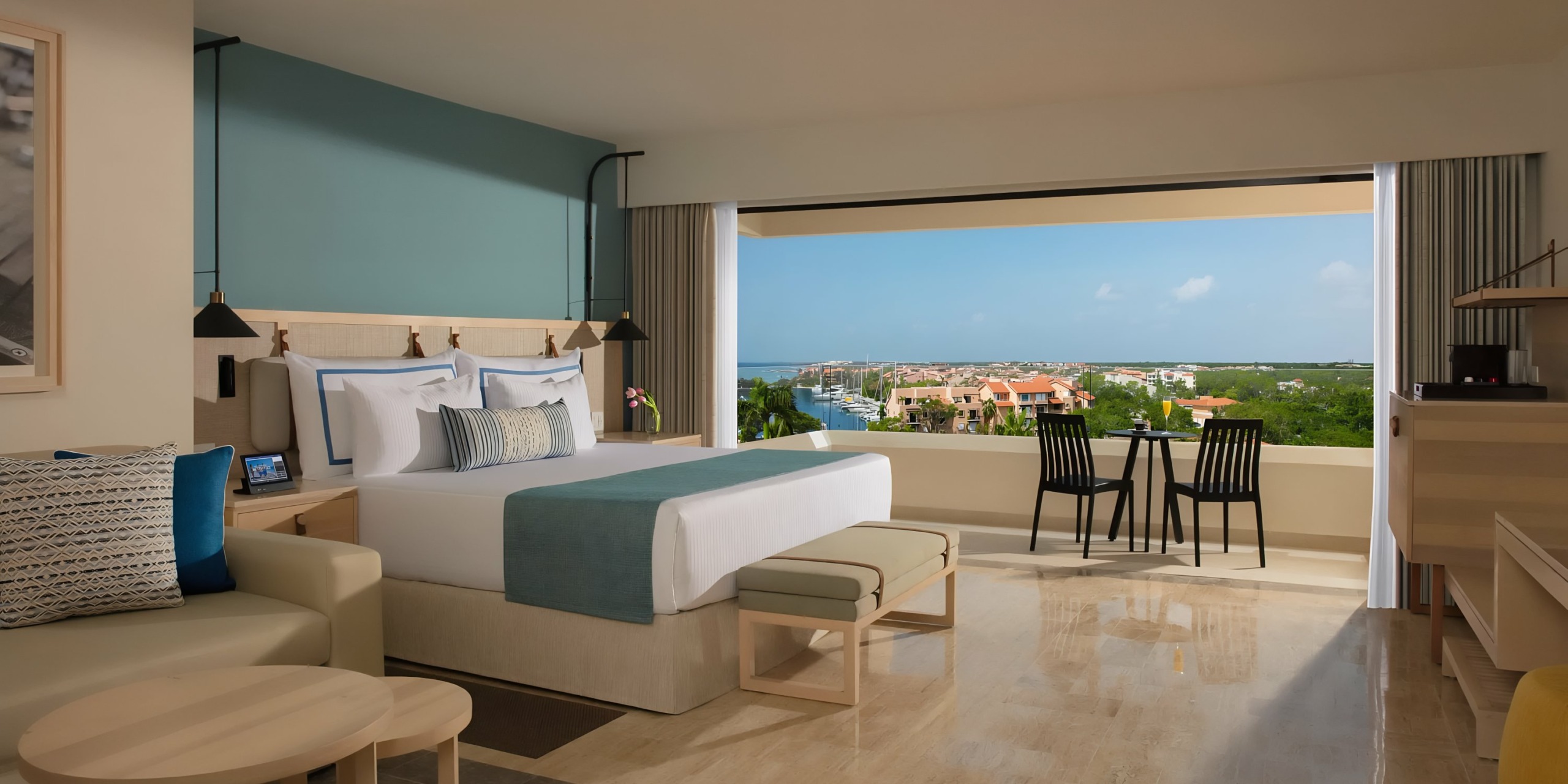 A hotel room with a Dreams Aventuras Riviera Maya bed, modern furnishings, and a large window offering a view of a coastal landscape.