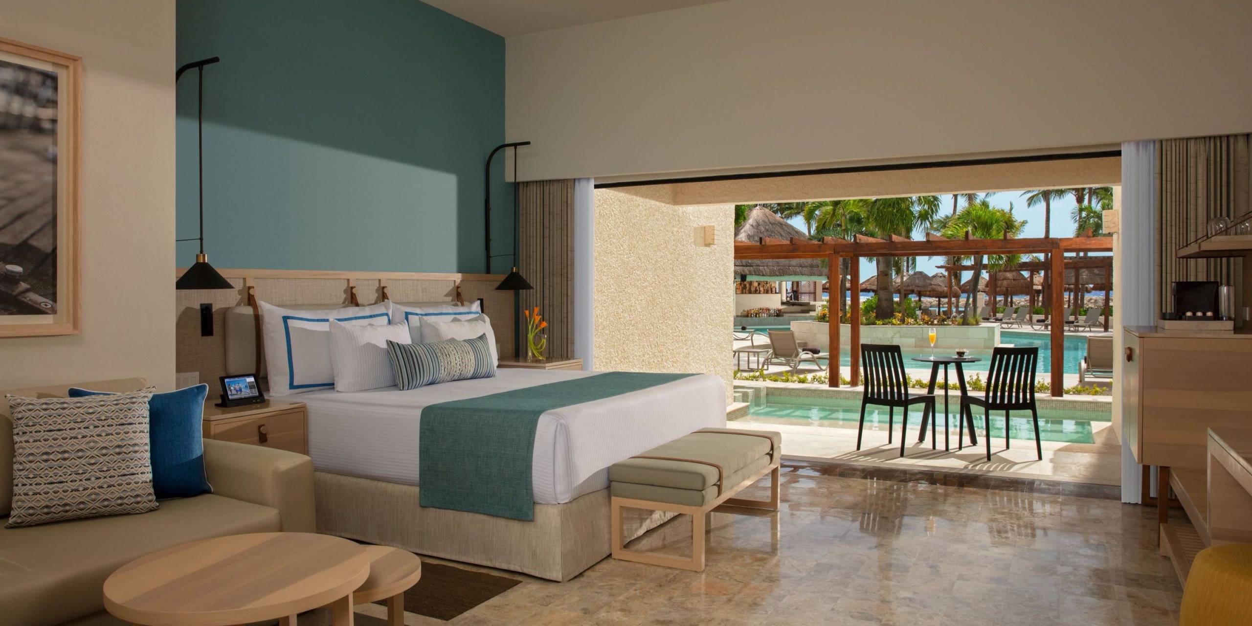 The Dreams Aventuras Riviera Maya offers a hotel room that includes a comfortable bed, a seating area, and an outdoor patio overlooking the pool. The patio is furnished with a table and chairs, and the room's decor features soothing teal and beige tones.