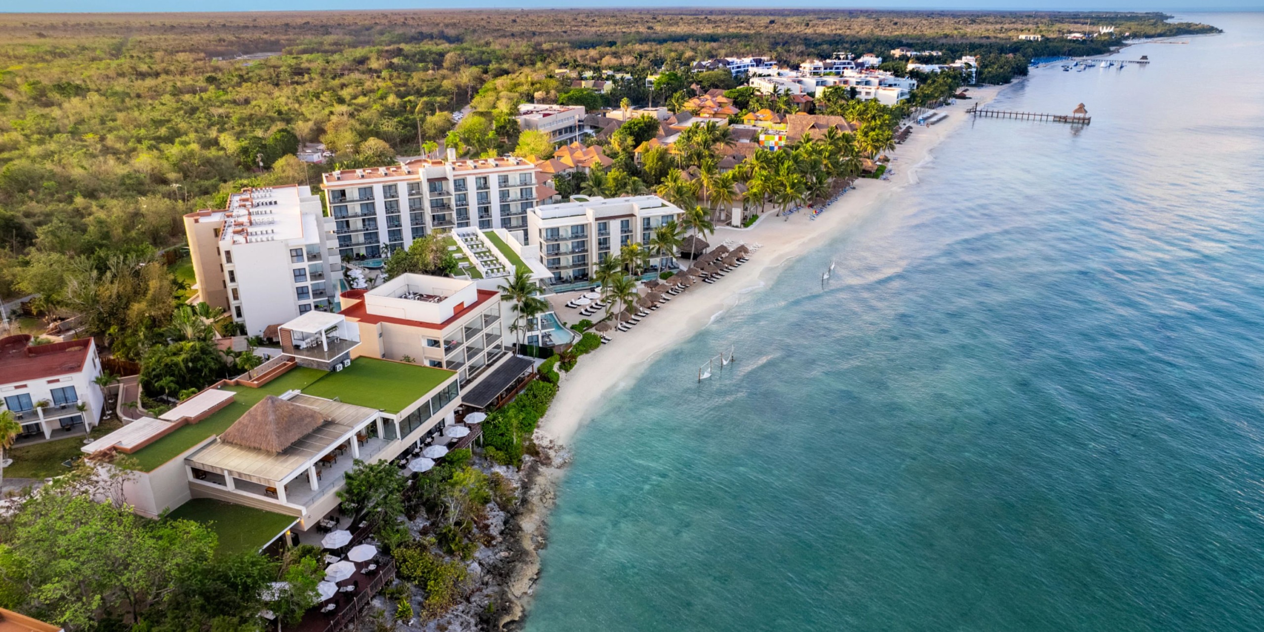 Aerial view of Dreams Cozumel Cape, a coastal resort featuring modern buildings, a sandy beach, palm trees, and clear blue water extending to the horizon.