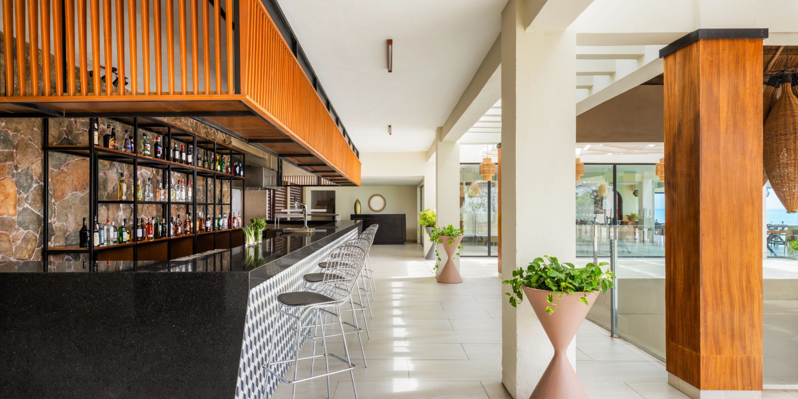 A contemporary bar featuring the Dreams Cozumel Cape with a sleek black countertop, high metal stools, and a stone accent wall. Large planters along with abundant natural light from the windows contribute to an inviting ambiance.