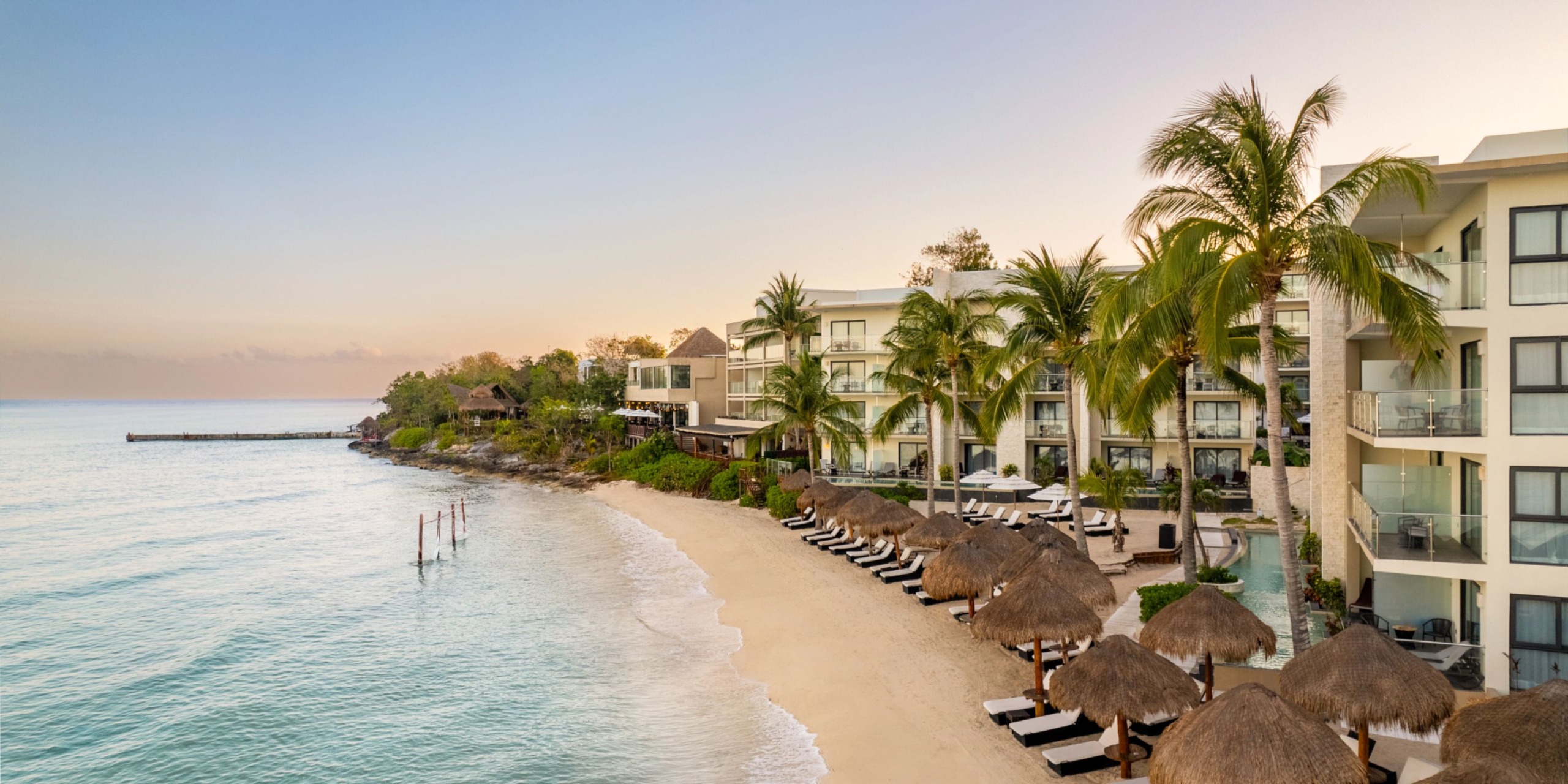 Dreams Cozumel Cape, a beachfront resort featuring multiple buildings, palm trees, and a row of thatched umbrellas along its sandy shore beside the serene sea at sunset.