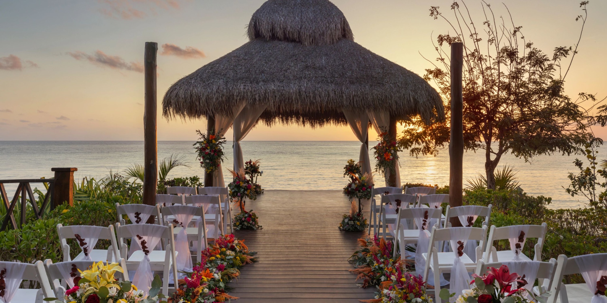 Experience the ultimate outdoor wedding setup at Dreams Cozumel Cape with a thatched gazebo overlooking the ocean at sunset, featuring rows of white chairs and vibrant floral arrangements lining the aisle.