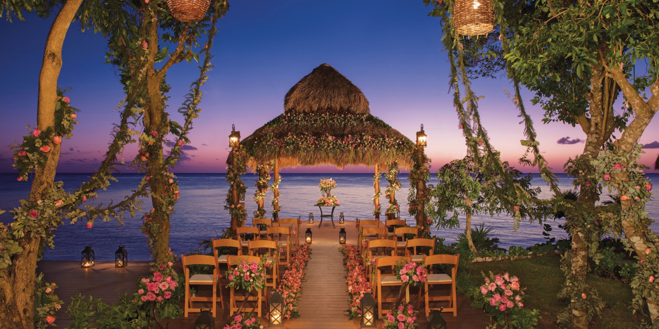 Dreams Cozumel Cape offers a stunning beachfront wedding setup featuring a floral-covered gazebo, wooden chairs beautifully adorned with flowers, and elegant lanterns, all set against a breathtaking sunset backdrop.