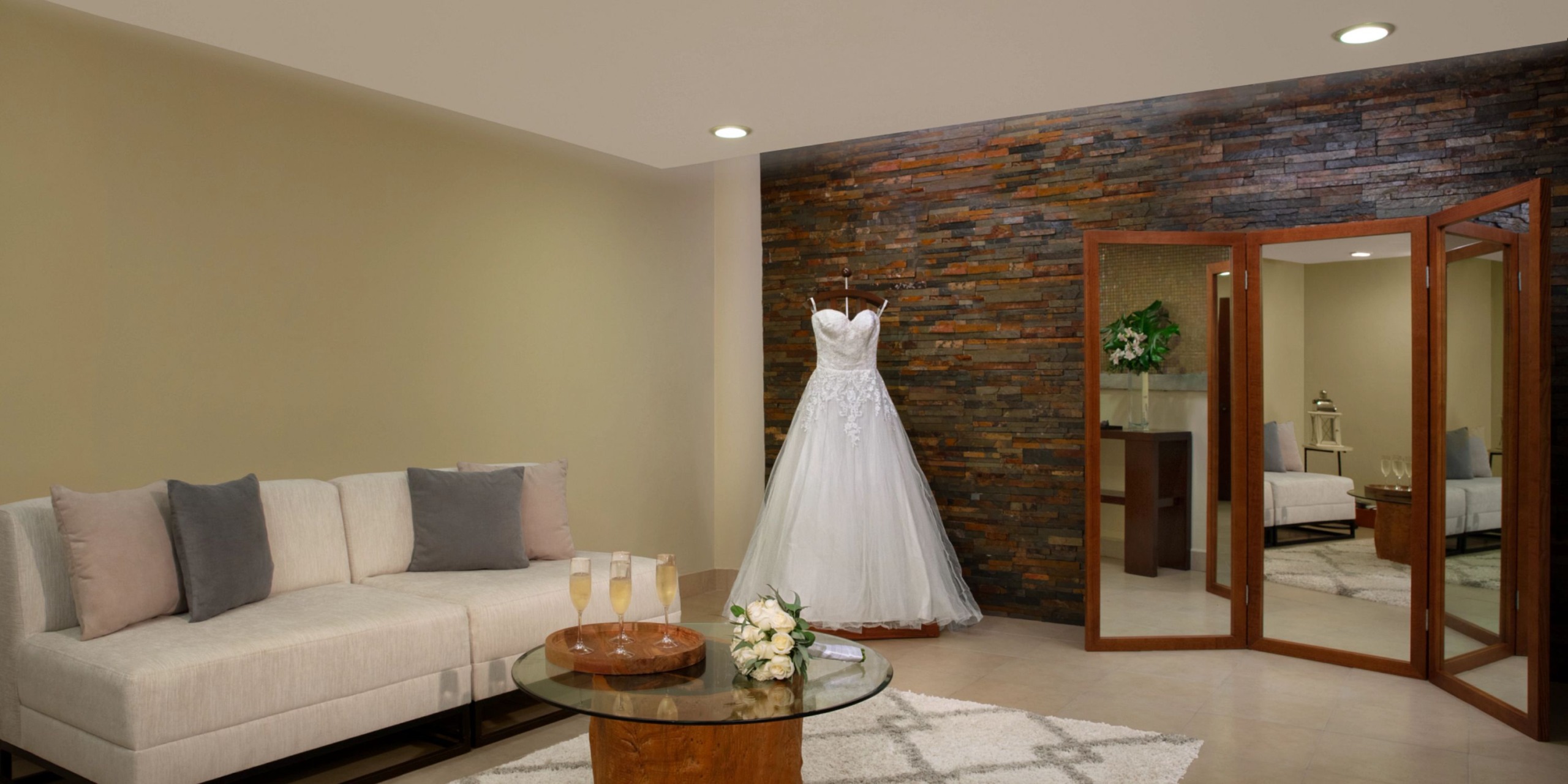 The Dreams Cozumel Cape bridal gown is elegantly displayed on a mannequin in a room featuring a stone accent wall, complemented by a three-panel mirror, a cream sofa, and a round glass table adorned with champagne flutes and a bouquet.