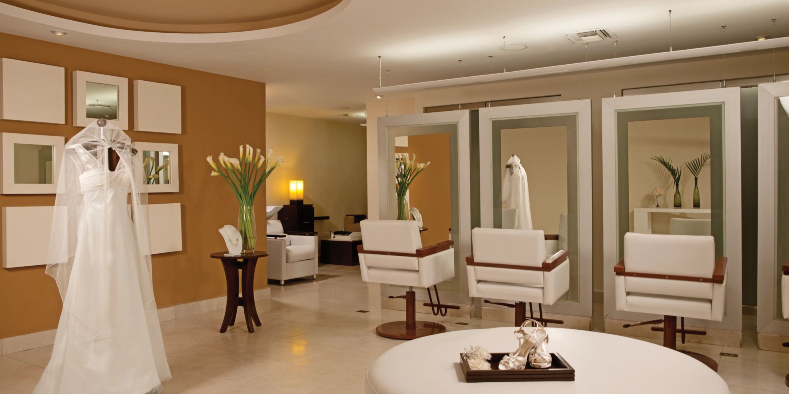 The interior of this contemporary bridal boutique features exquisite Dreams Cozumel Capes, complemented by white chairs, mirrors, and floral arrangements.