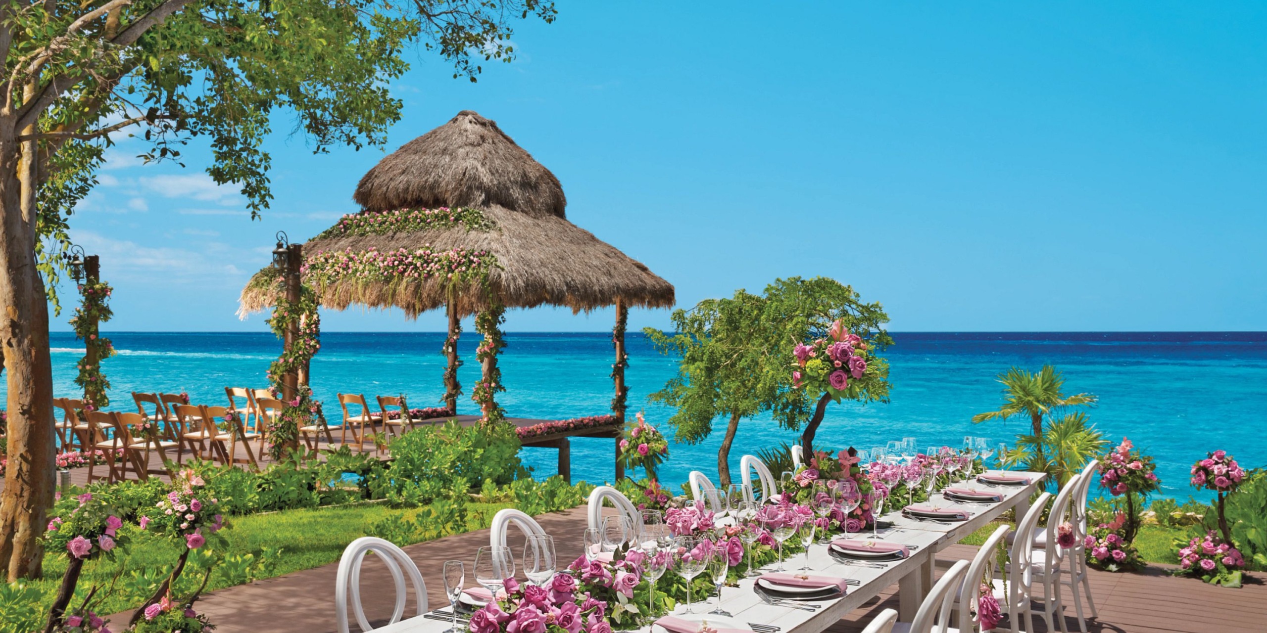 Enjoy a dining experience at Dreams Cozumel Cape, featuring a long table adorned with pink flowers beneath a thatched pergola, all set against the backdrop of crystal-clear blue waters.