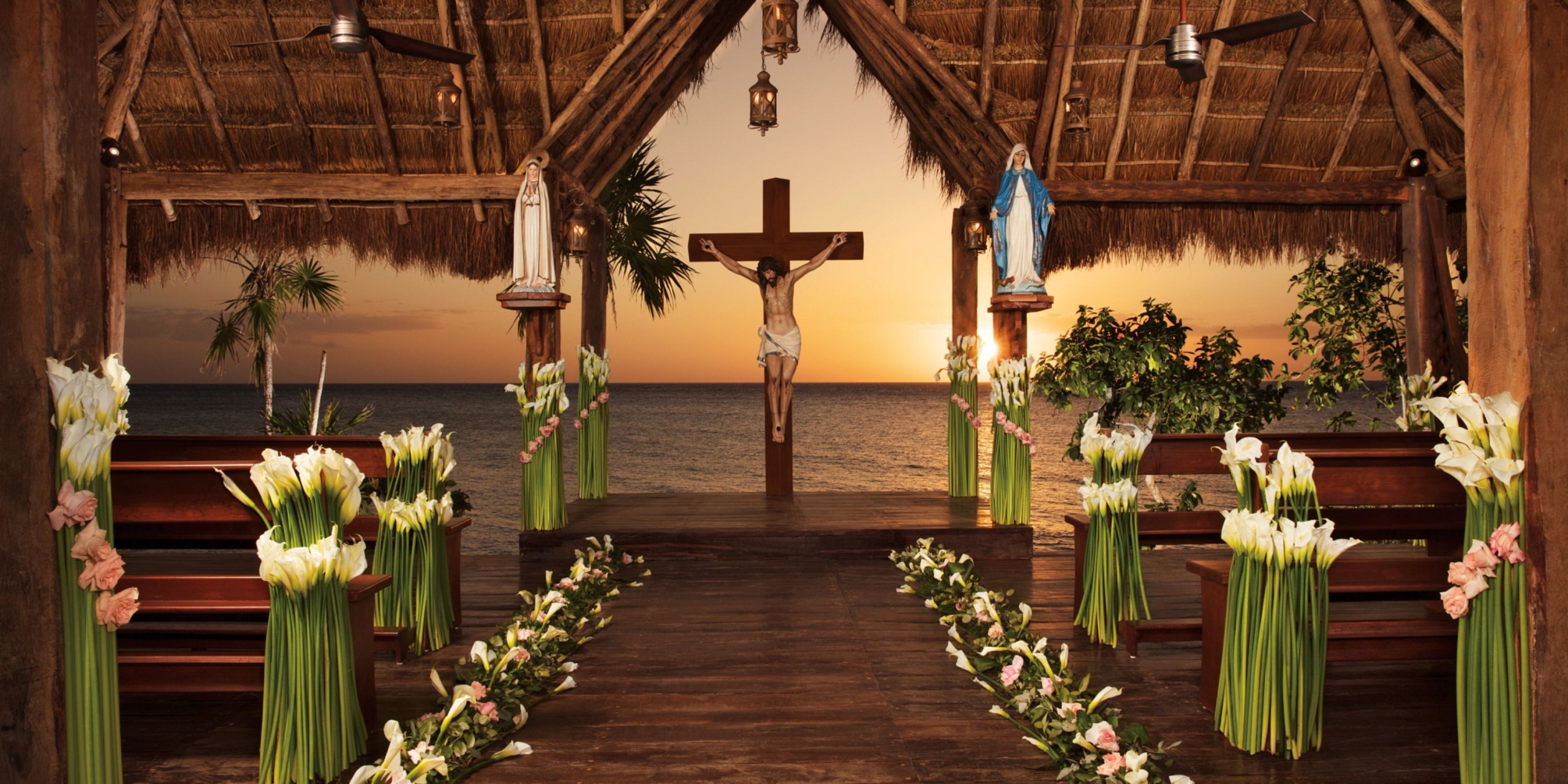 The Dreams Cozumel Cape offers an outdoor chapel featuring a thatched roof and floral decorations, overlooking the ocean at sunset with a crucifix centered at the altar.