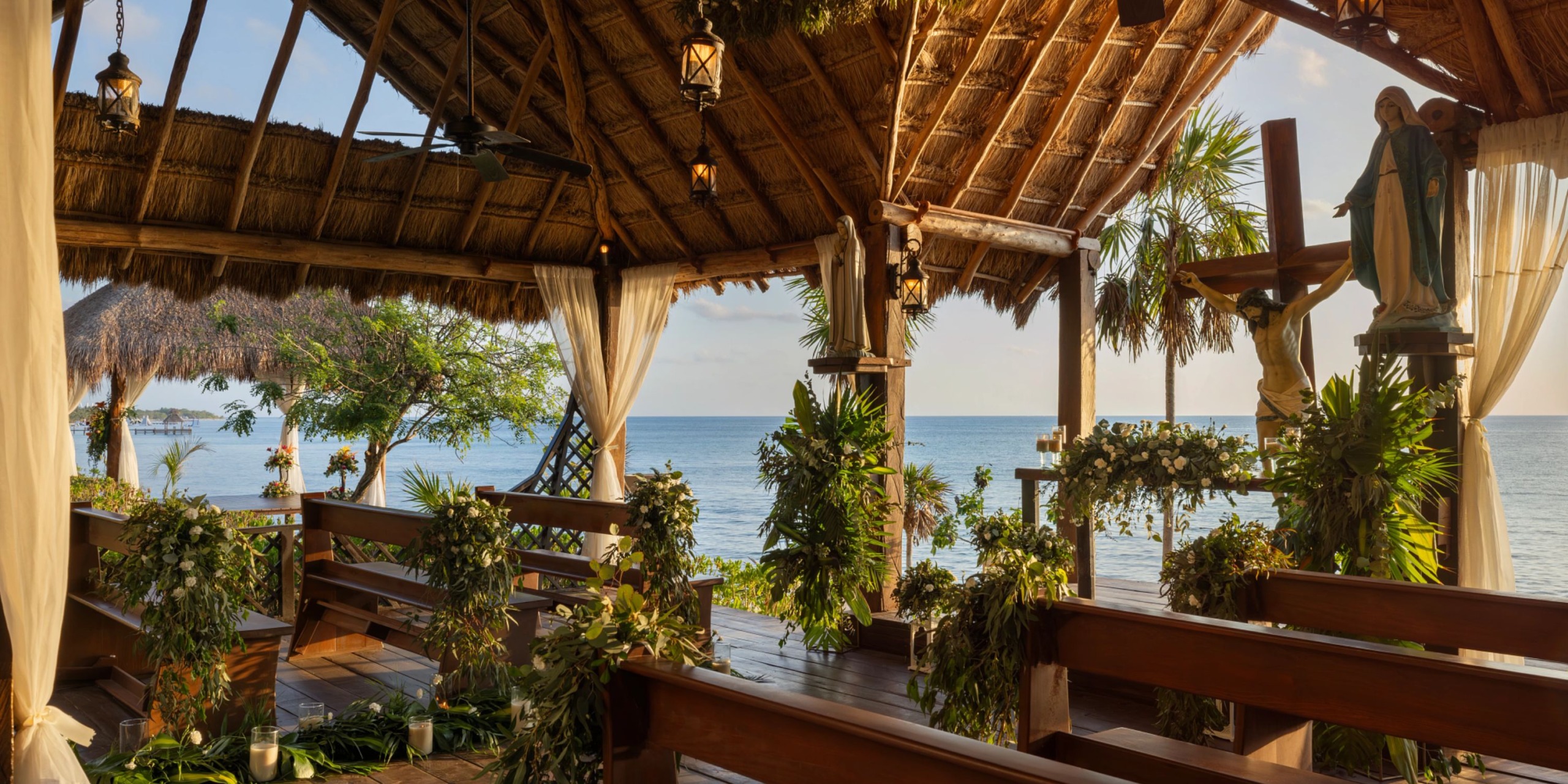 Step into the Dreams Cozumel Cape, a charming beachside chapel featuring wooden benches and a thatched roof, all set against an ocean view. The space is beautifully adorned with greenery and floral arrangements.