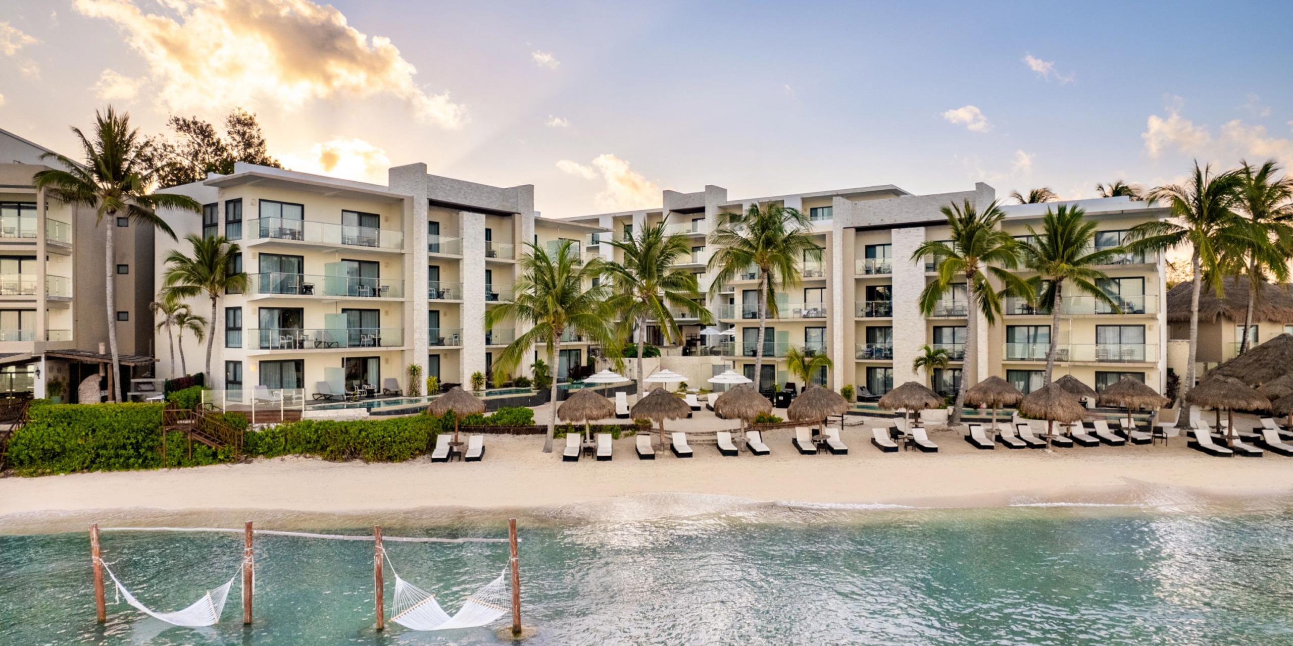 Dreams Cozumel Cape, a beachfront resort featuring comfy loungers, swaying palm trees, and serene hammocks over the water. In the background, elegant four-story white buildings stand under a partly cloudy sky.